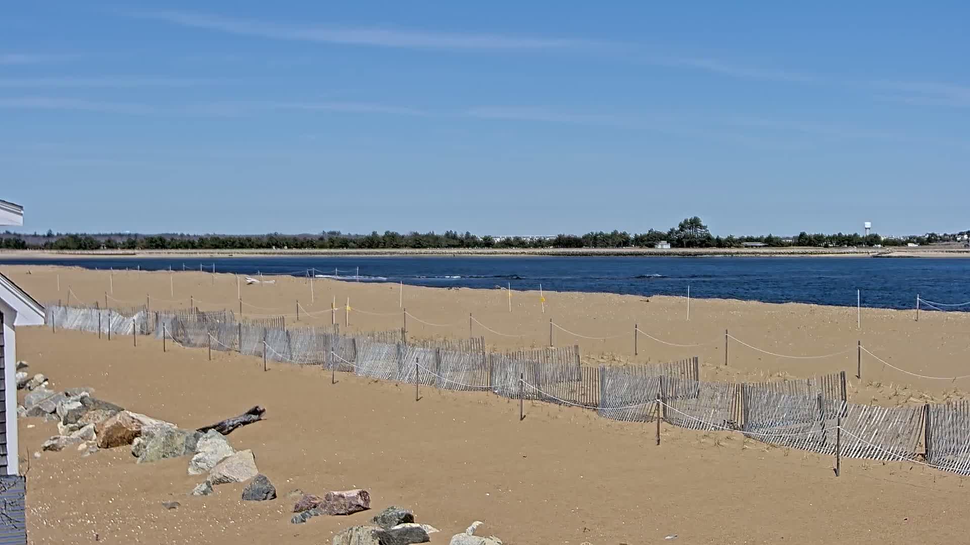 A sandy beach with a wooden fence and rocks, under a clear blue sky, overlooks a calm body of water and a tree line on the opposite shore.