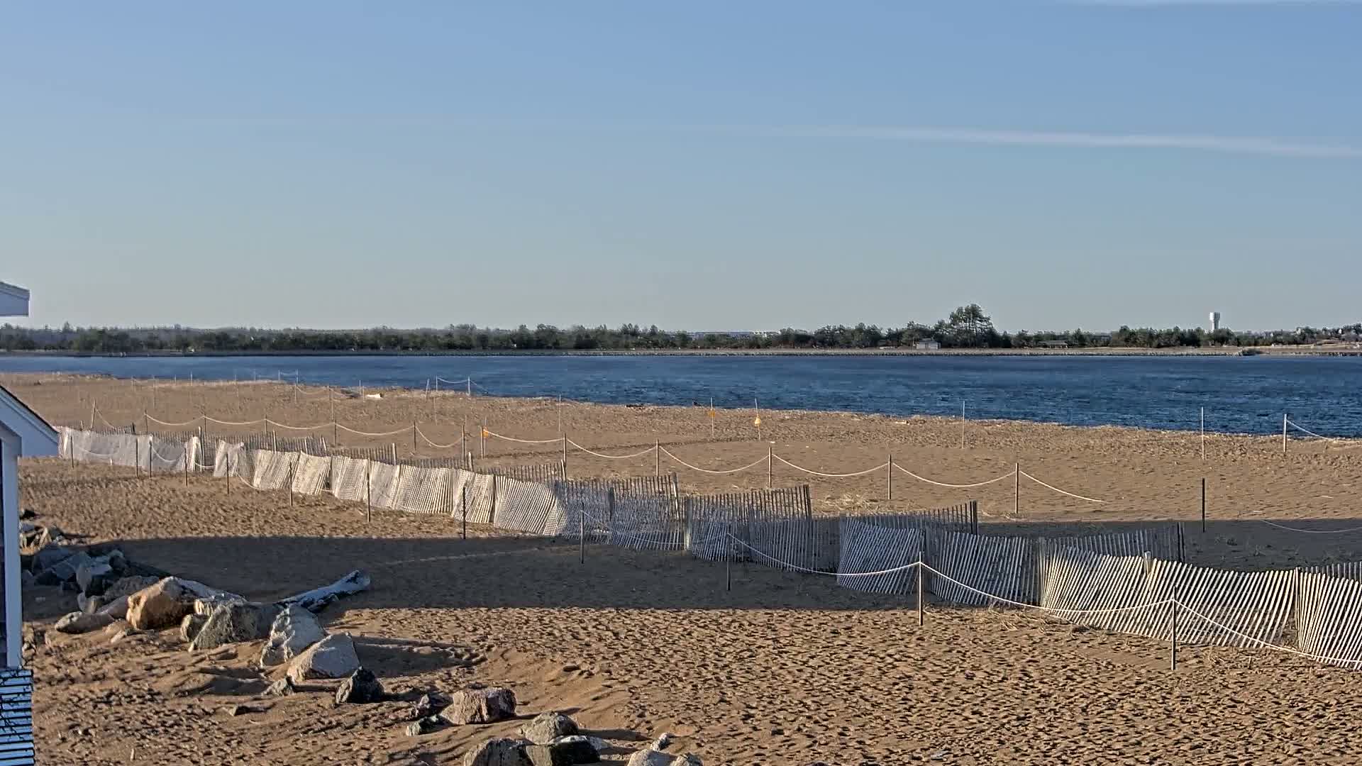A sandy beach with short, white fences stretches towards a calm body of water under a clear, sunny sky.