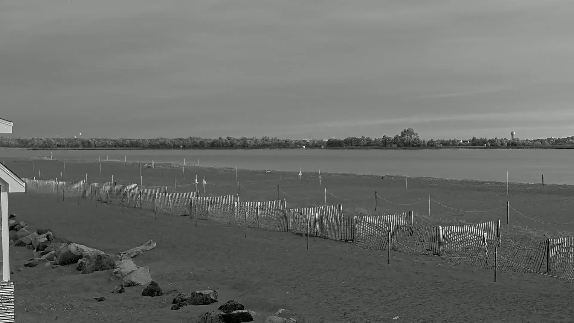 A sandy beach with a low wooden fence stretches along a calm body of water under a cloudy sky, with a distant treeline visible on the opposite shore.
