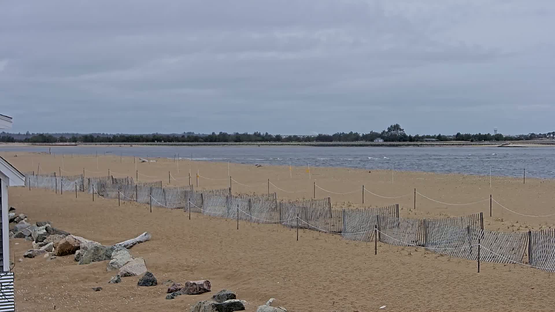 A sandy beach with a short wooden fence stretches along the shoreline, under a cloudy sky, overlooking calm water and a distant tree line.