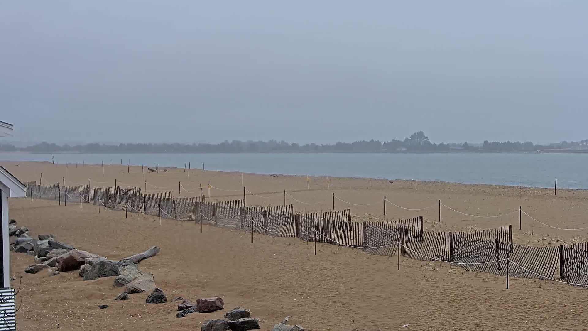A sandy beach with a low wooden fence stretches towards a calm body of water under an overcast sky.