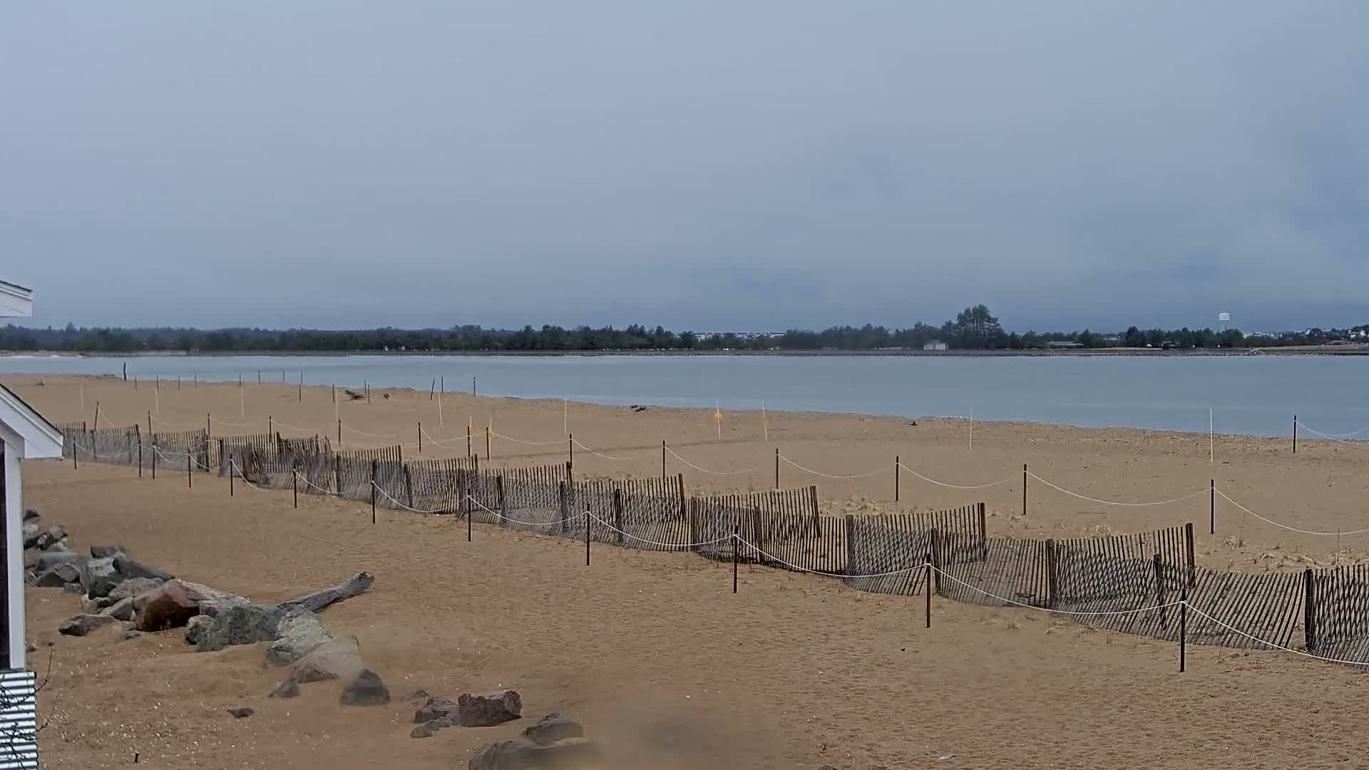 A sandy beach with a low wooden fence stretches along a calm, gray body of water under an overcast sky.