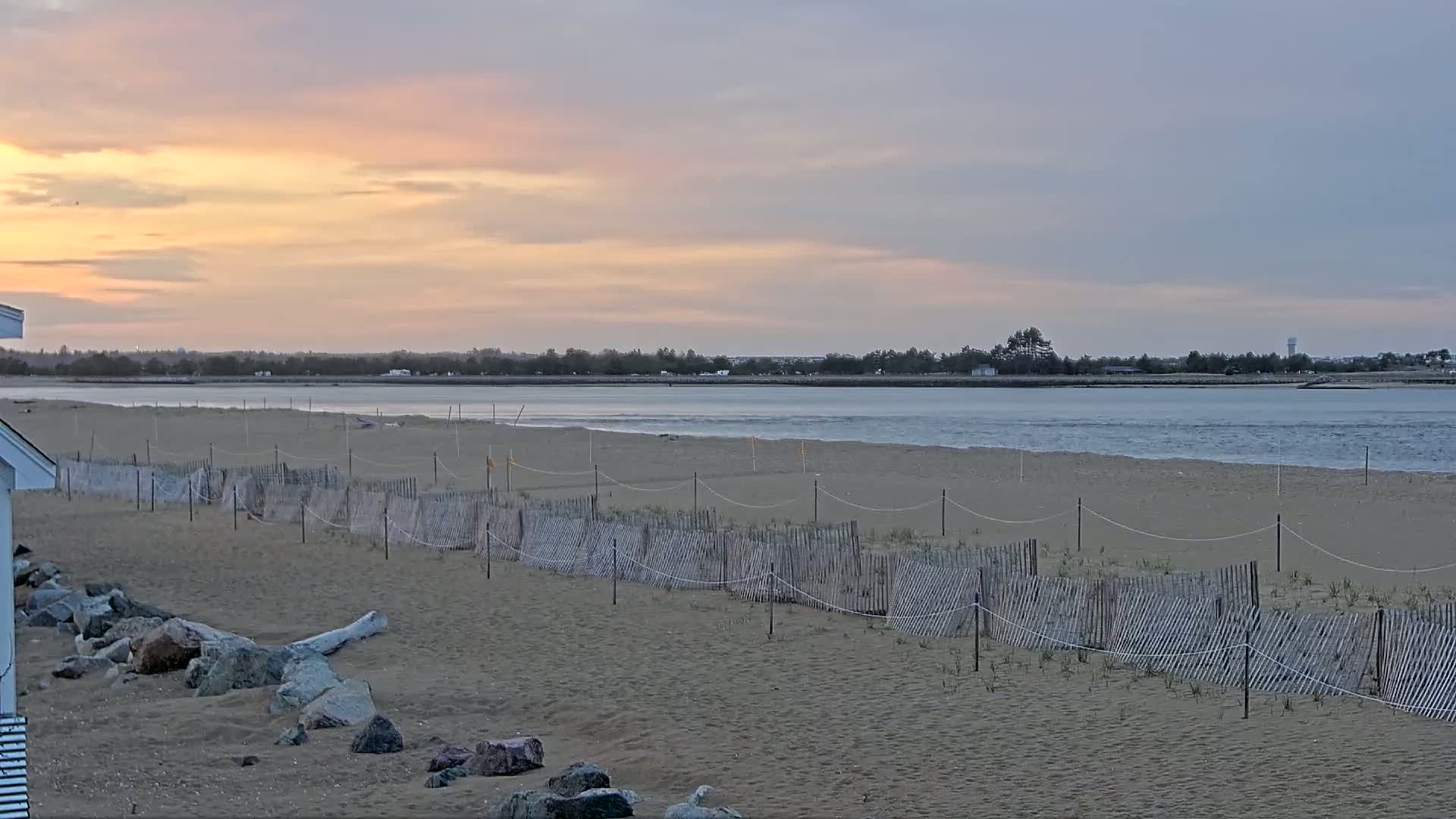 A sandy beach with a low wooden fence and calm water under a pastel sunrise sky.