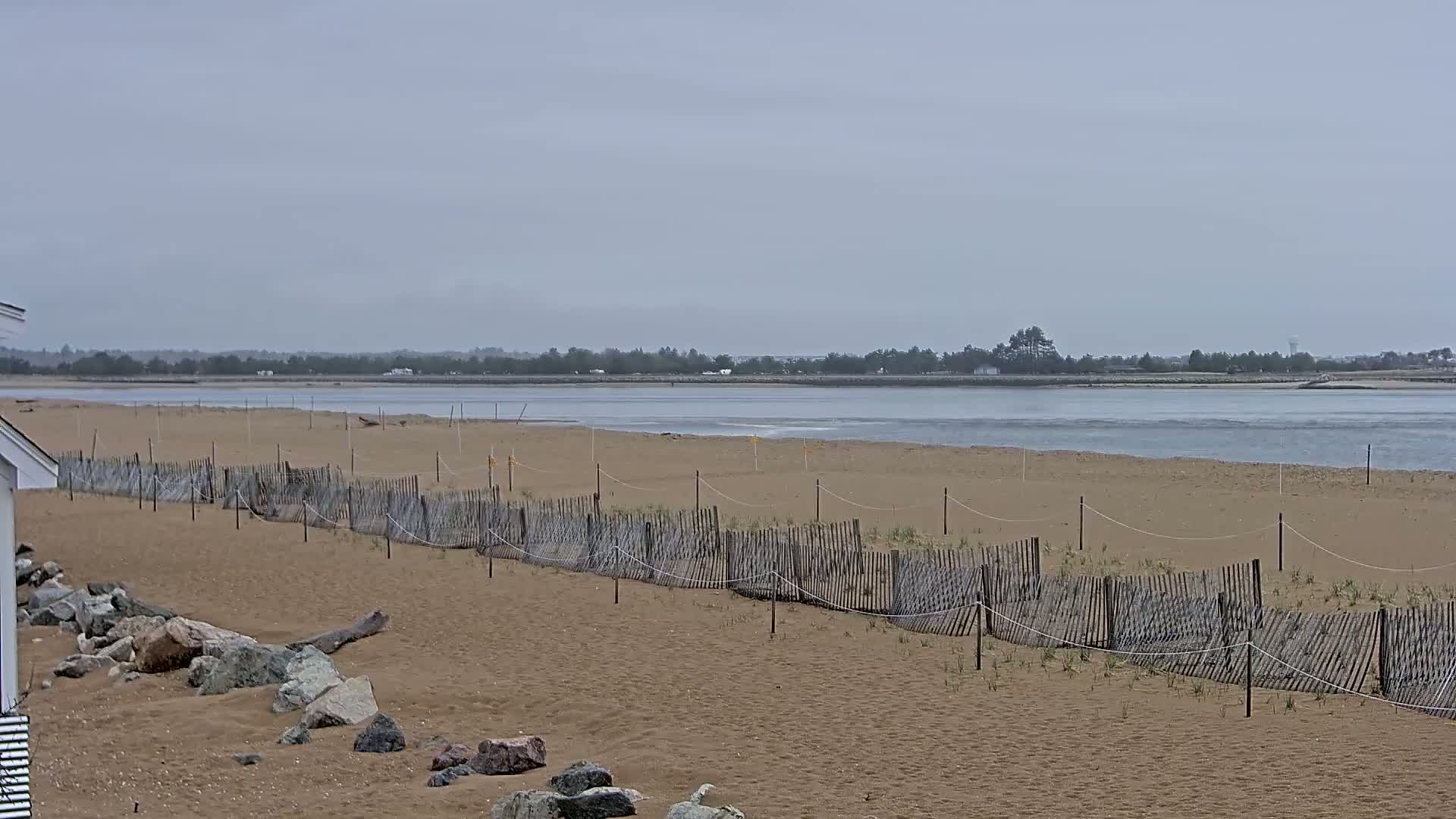 A sandy beach with a low wooden fence stretches towards a calm, gray-toned body of water under an overcast sky.