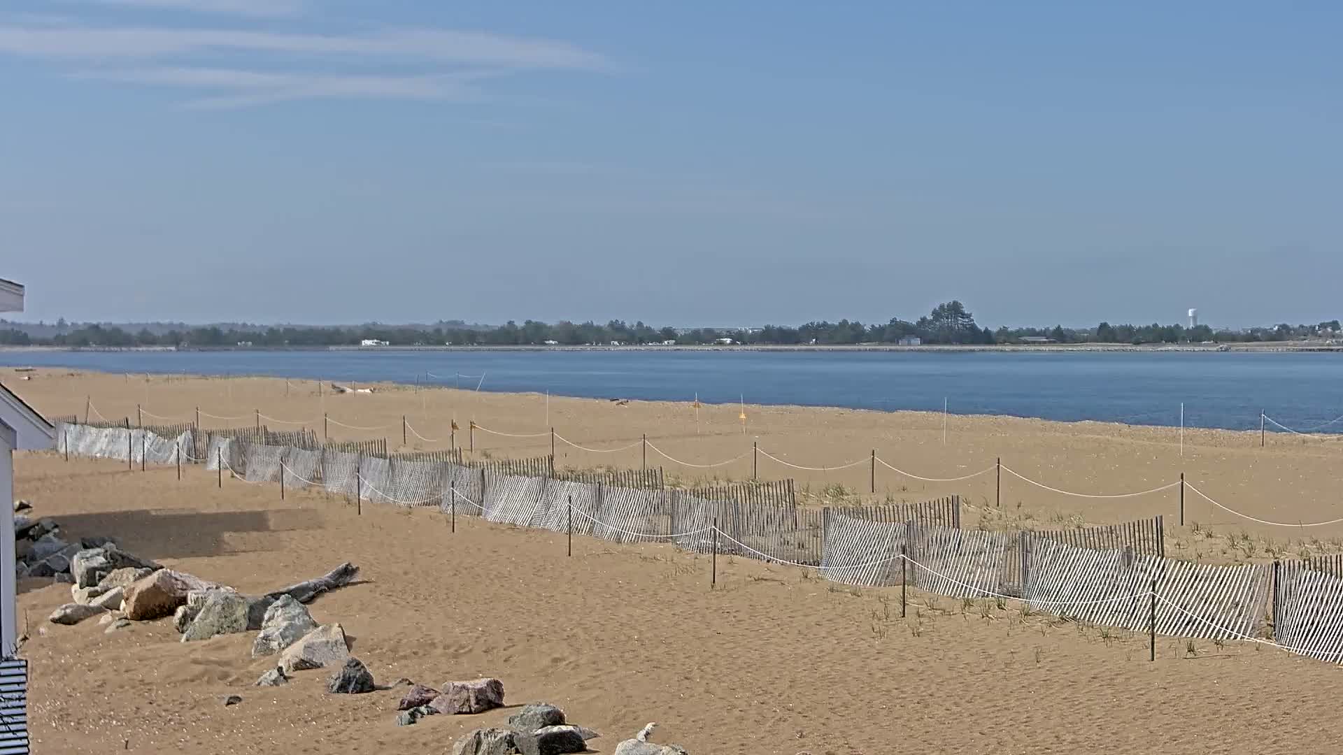 A sandy beach with a low wooden fence stretches along a calm body of water under a clear, sunny sky.