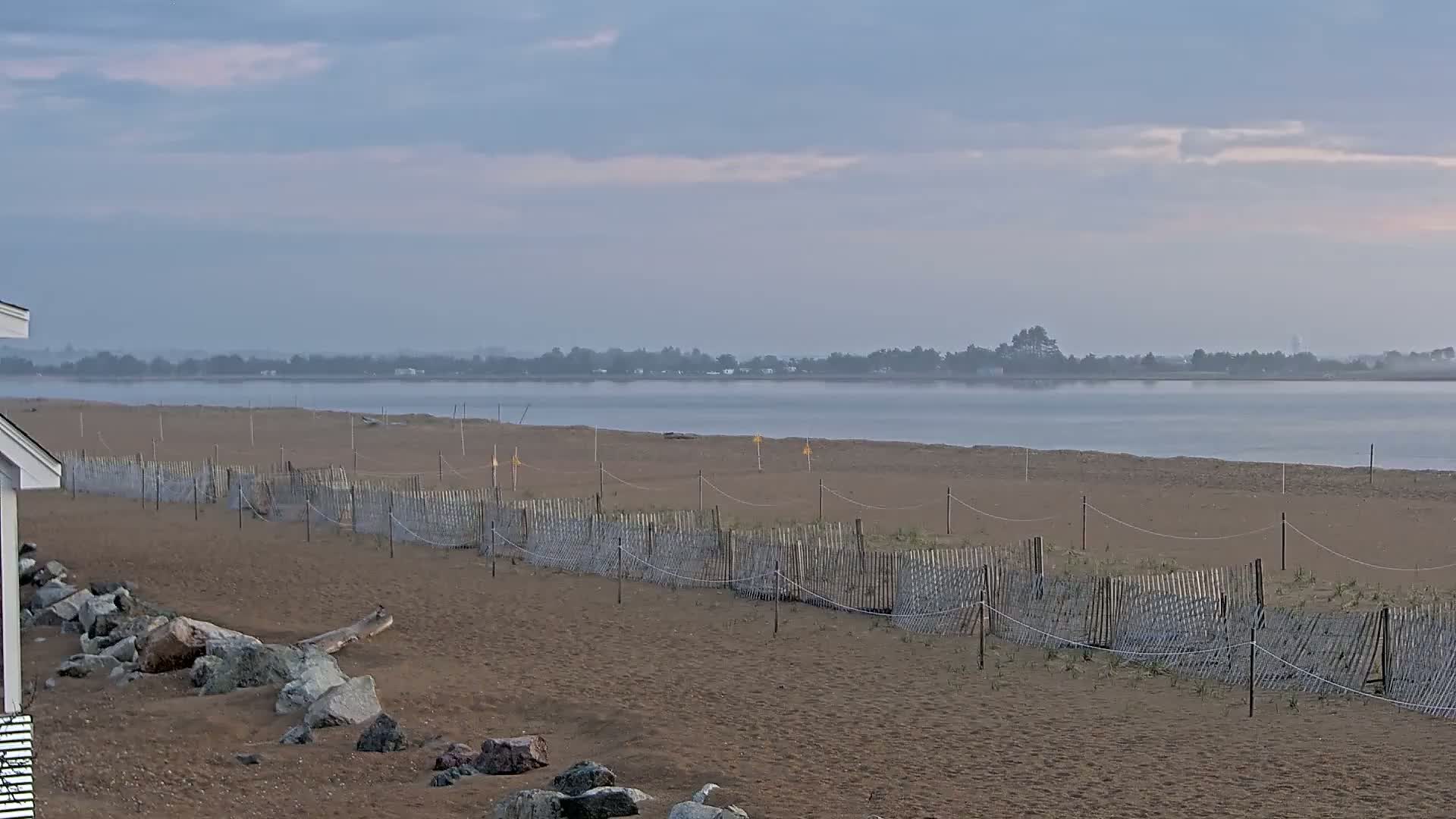 A sandy beach with a low wooden fence stretches along a calm body of water under a hazy sky.