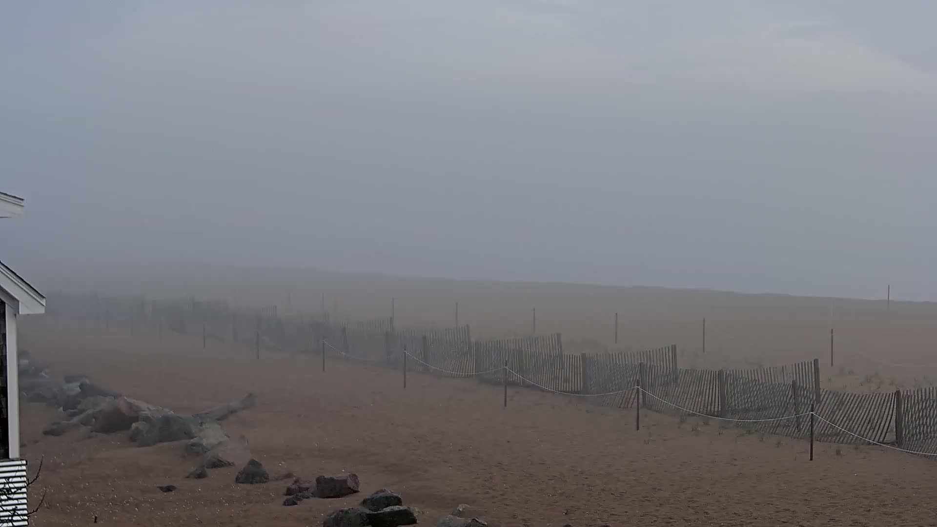 A sandy beach, partially obscured by fog, features a weathered wooden fence running along its edge, near a building.