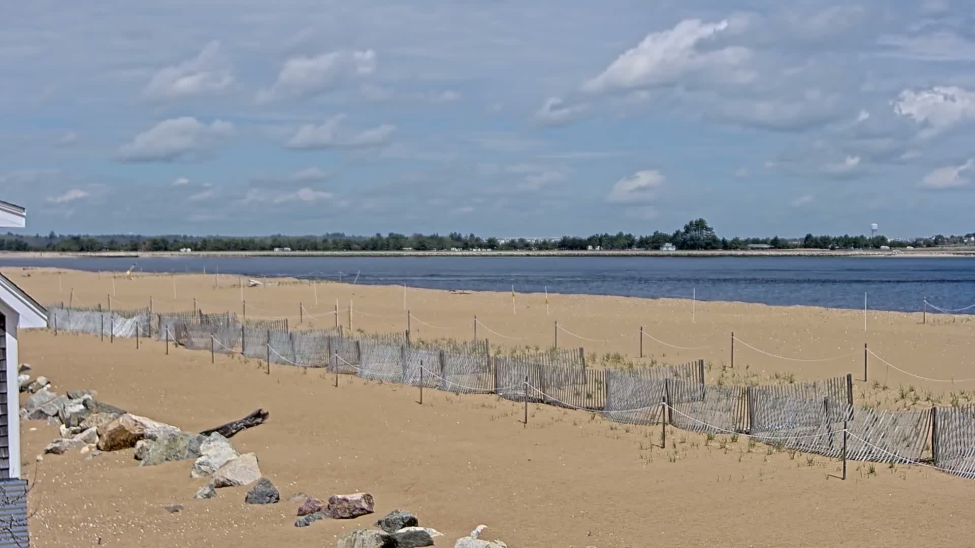 A sandy beach with a low wooden fence and calm water beyond it under a partly cloudy sky.