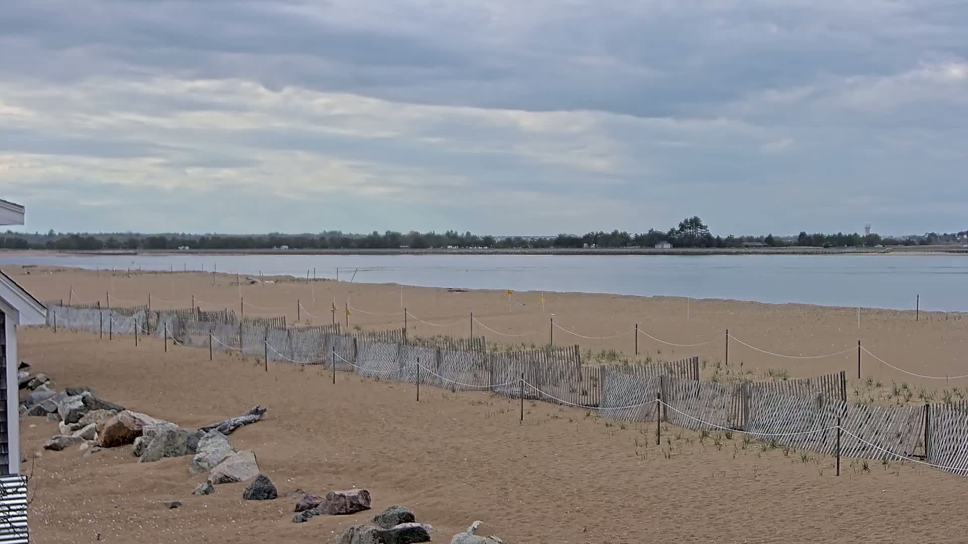 A sandy beach with a low wooden fence stretches towards a calm body of water under a cloudy sky.
