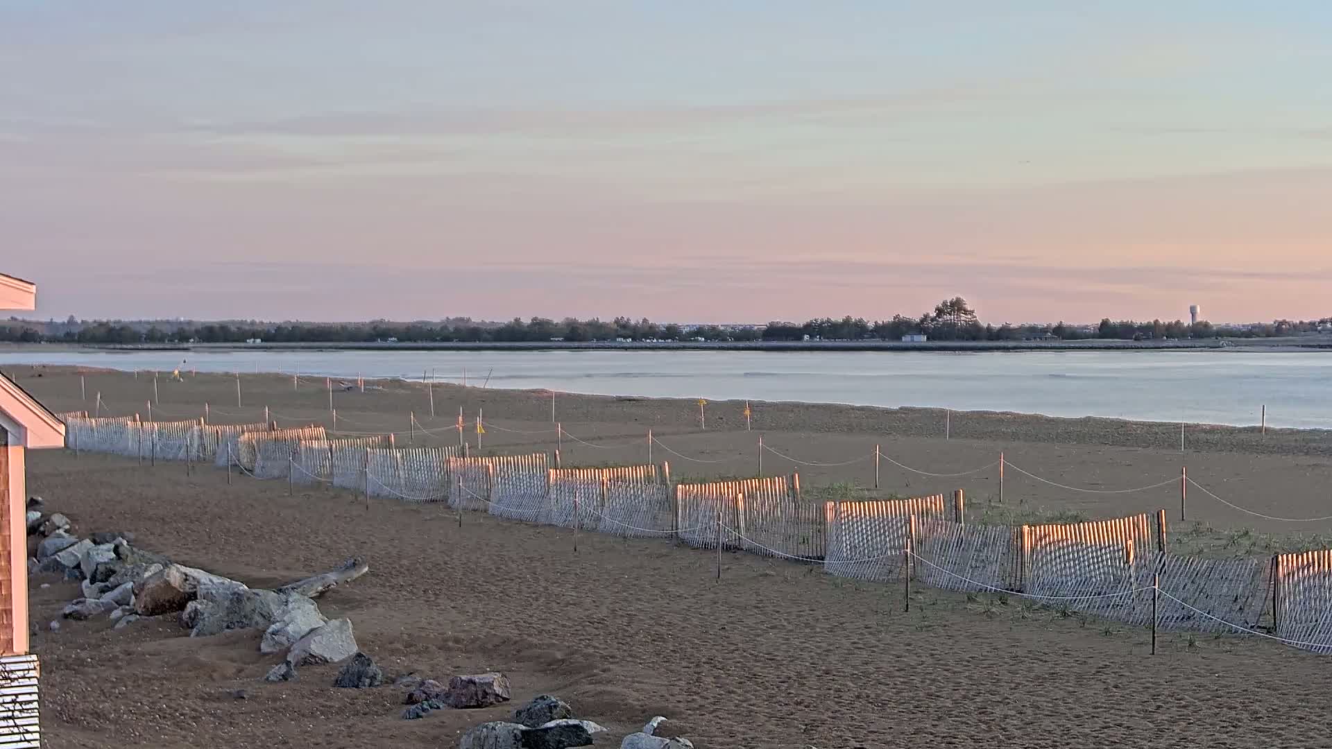 A sandy beach with a low wooden fence stretches toward a calm body of water under a pale pink sunset sky.
