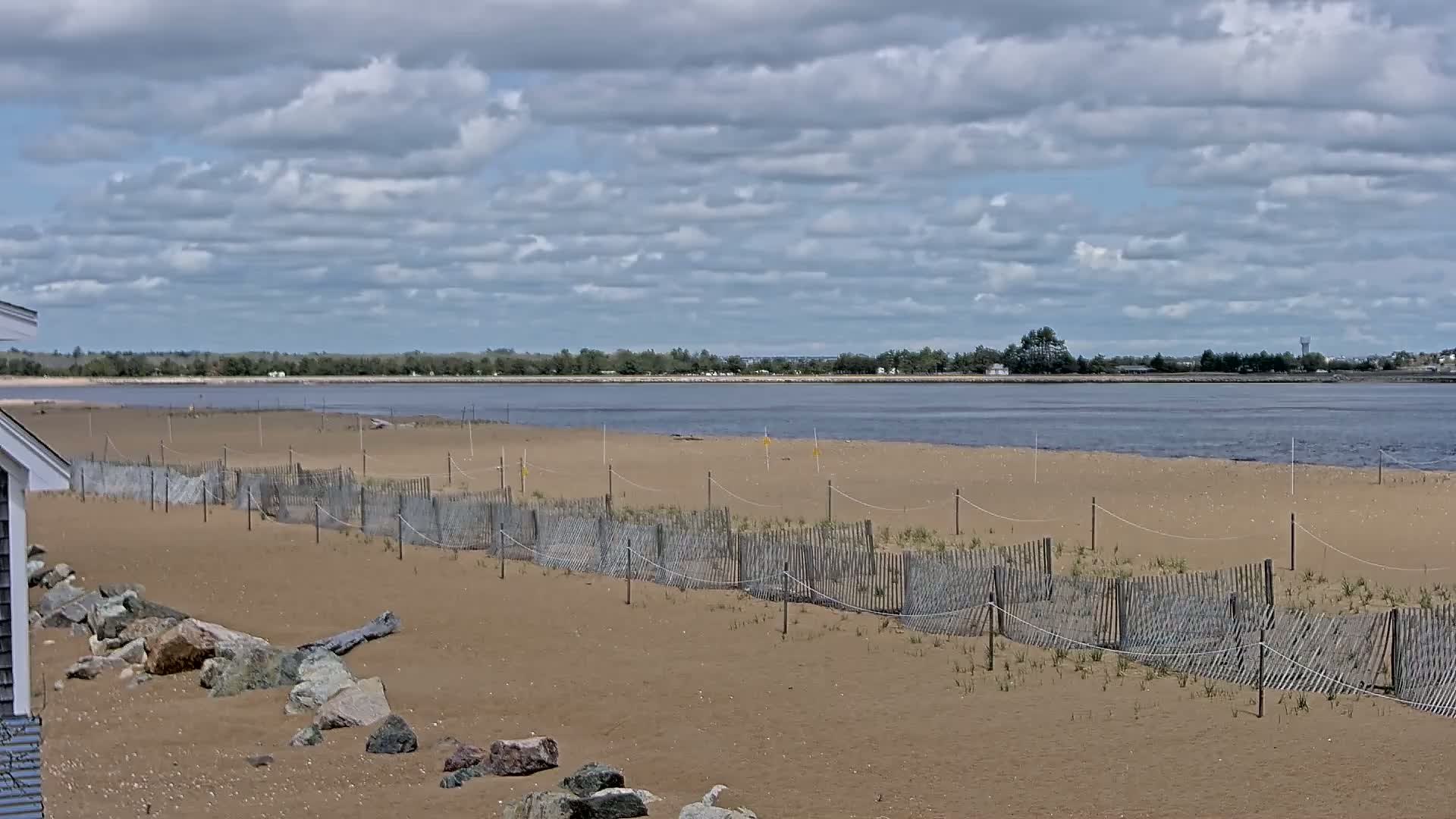 A sandy beach with a low wooden fence and scattered rocks stretches to a calm body of water under a partly cloudy sky.