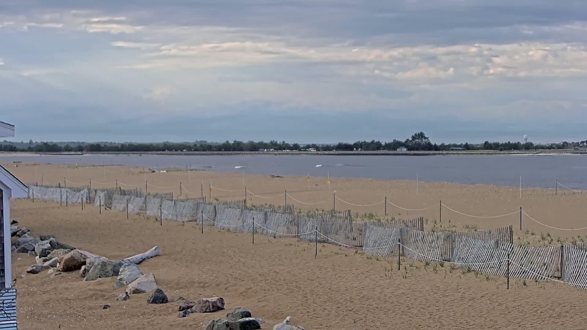 A sandy beach with a low wooden fence stretches towards a calm, grey-blue body of water under a cloudy sky.