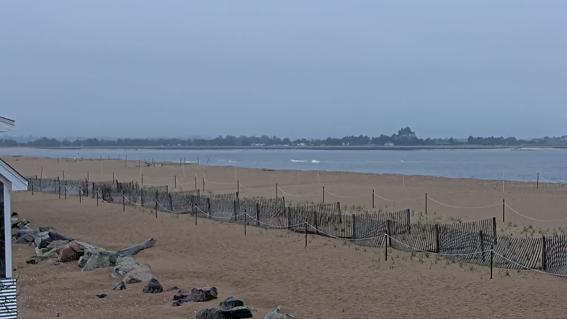 A sandy beach with a low wooden fence stretches toward a calm, gray body of water under an overcast sky.