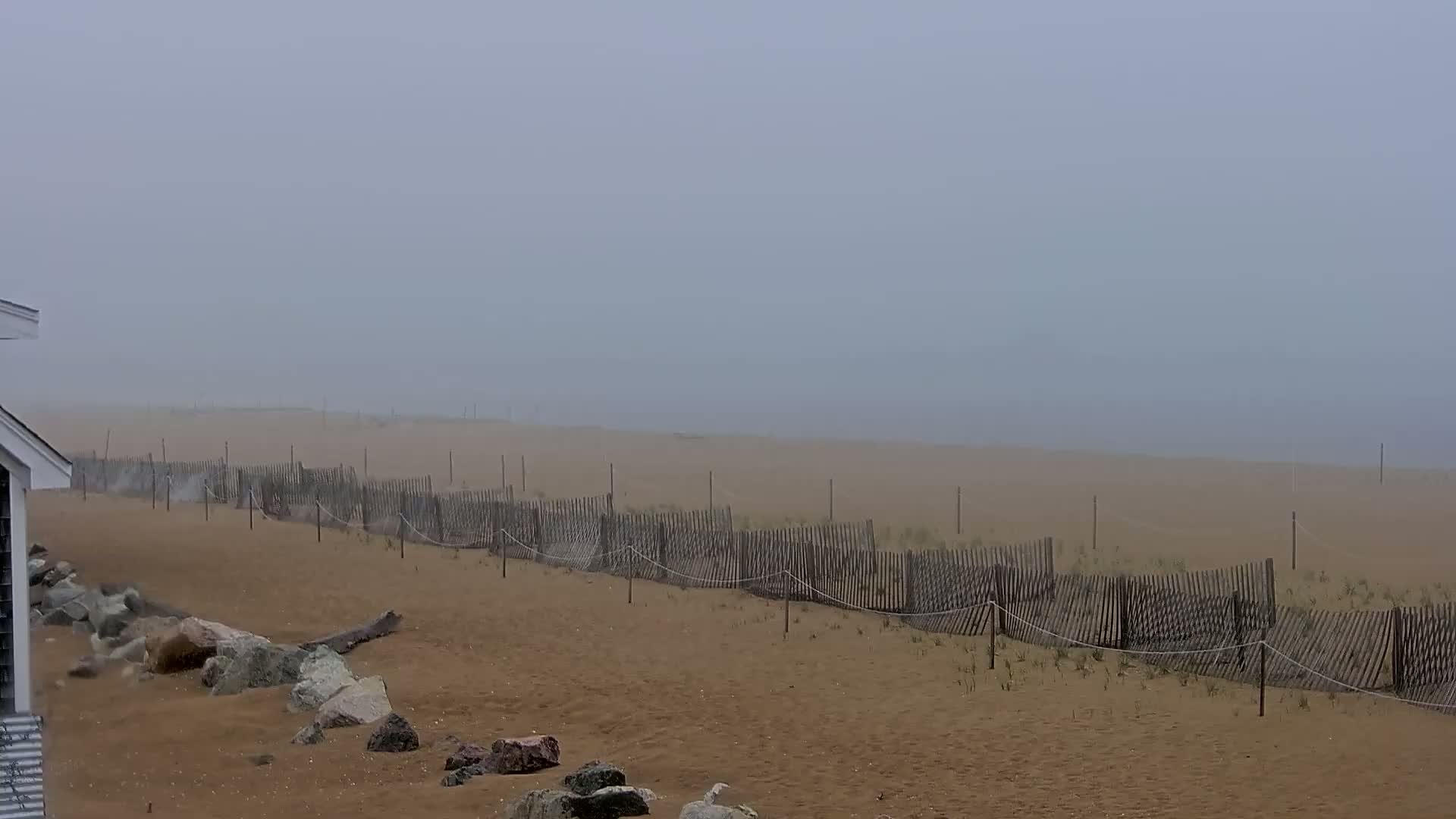 A sandy beach stretches into a foggy distance, behind a wooden fence, with rocks visible near a building in the foreground.