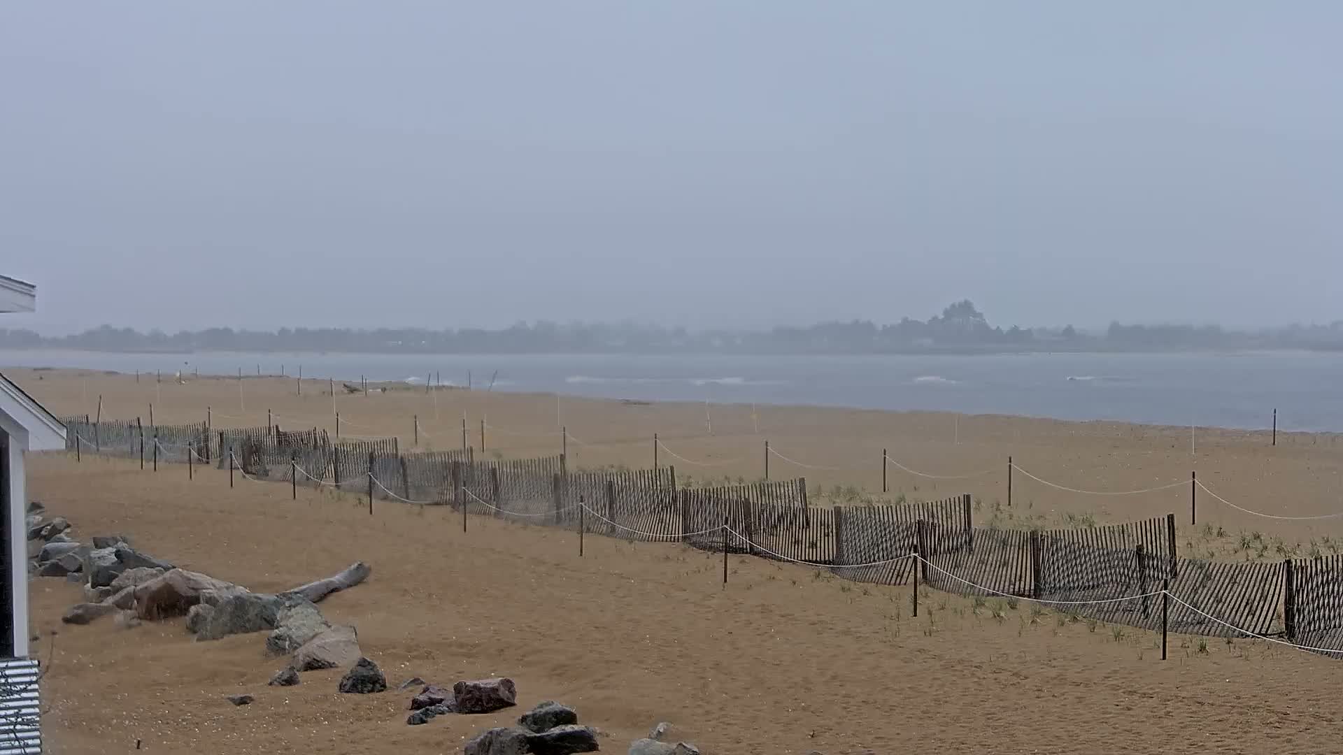 A sandy beach with a low wooden fence stretches along a calm, grey body of water under an overcast sky.