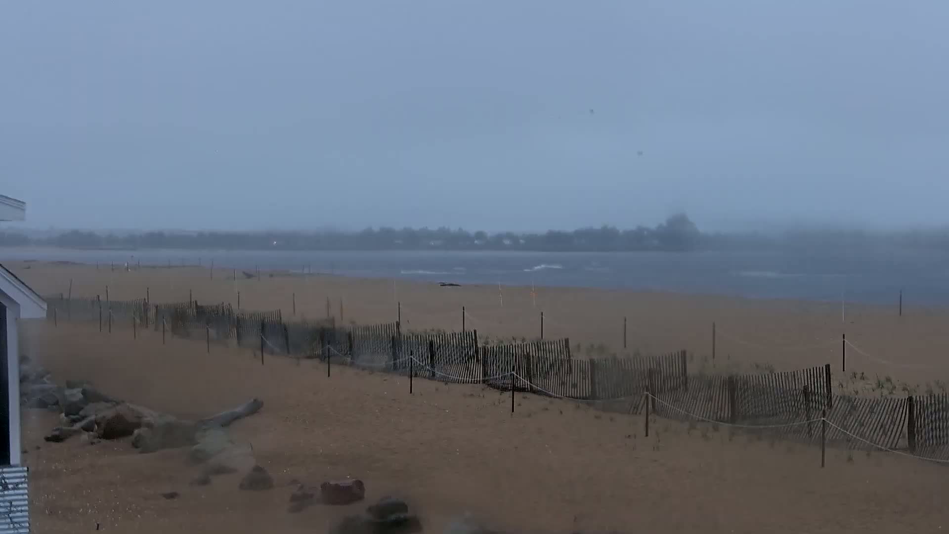A sandy beach with a fence is shown during a stormy, overcast day, with a body of water in the background.
