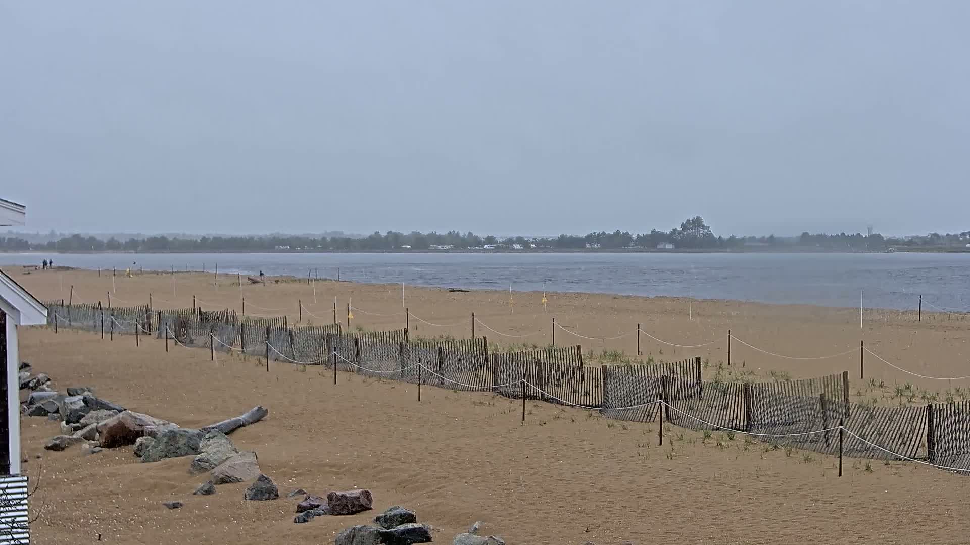 A sandy beach with a low wooden fence stretches towards a calm, grey body of water under an overcast sky.