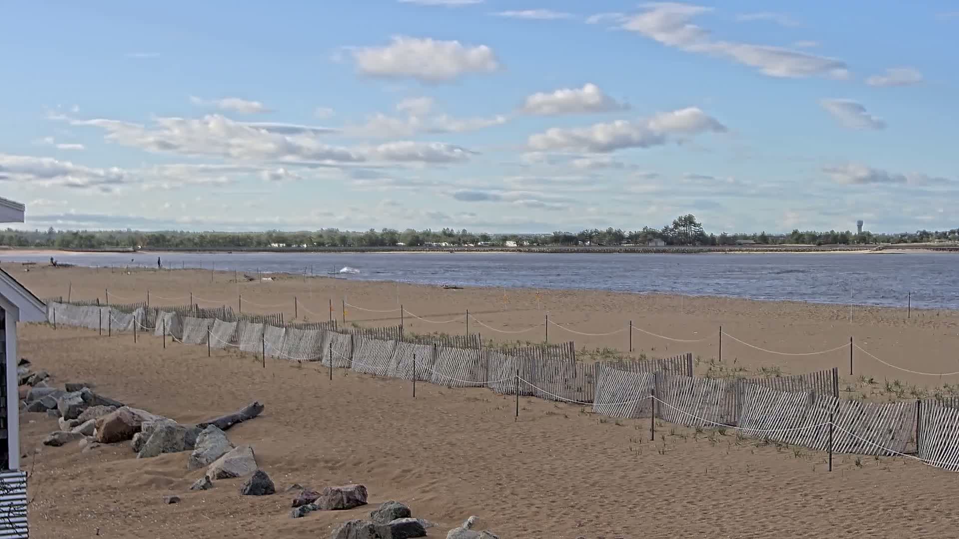 A sandy beach with a low wooden fence stretches toward a calm, somewhat choppy body of water under a partly cloudy sky.