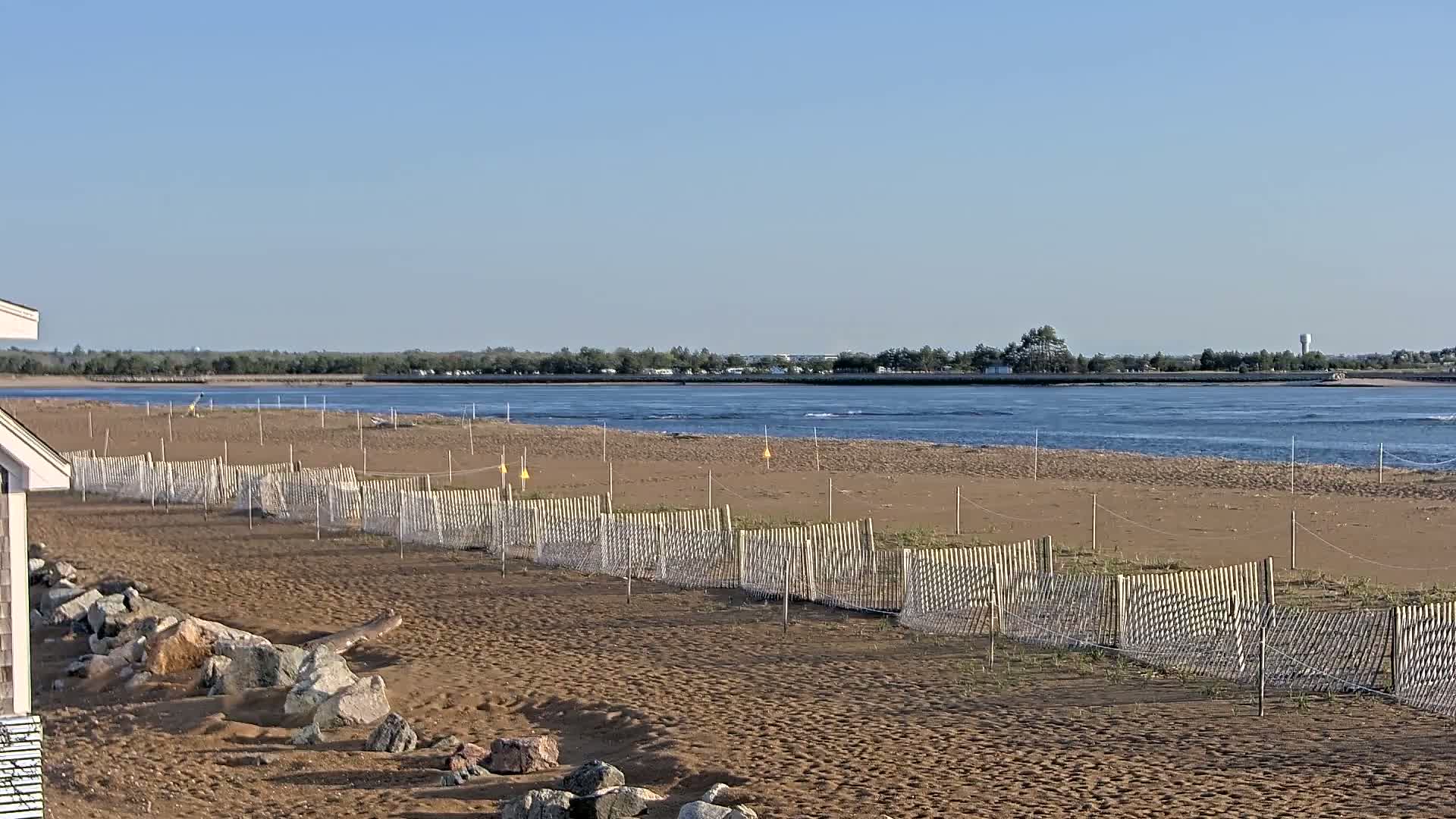 A sandy beach with low wooden fences and netting stretches towards a calm body of water under a clear blue sky.