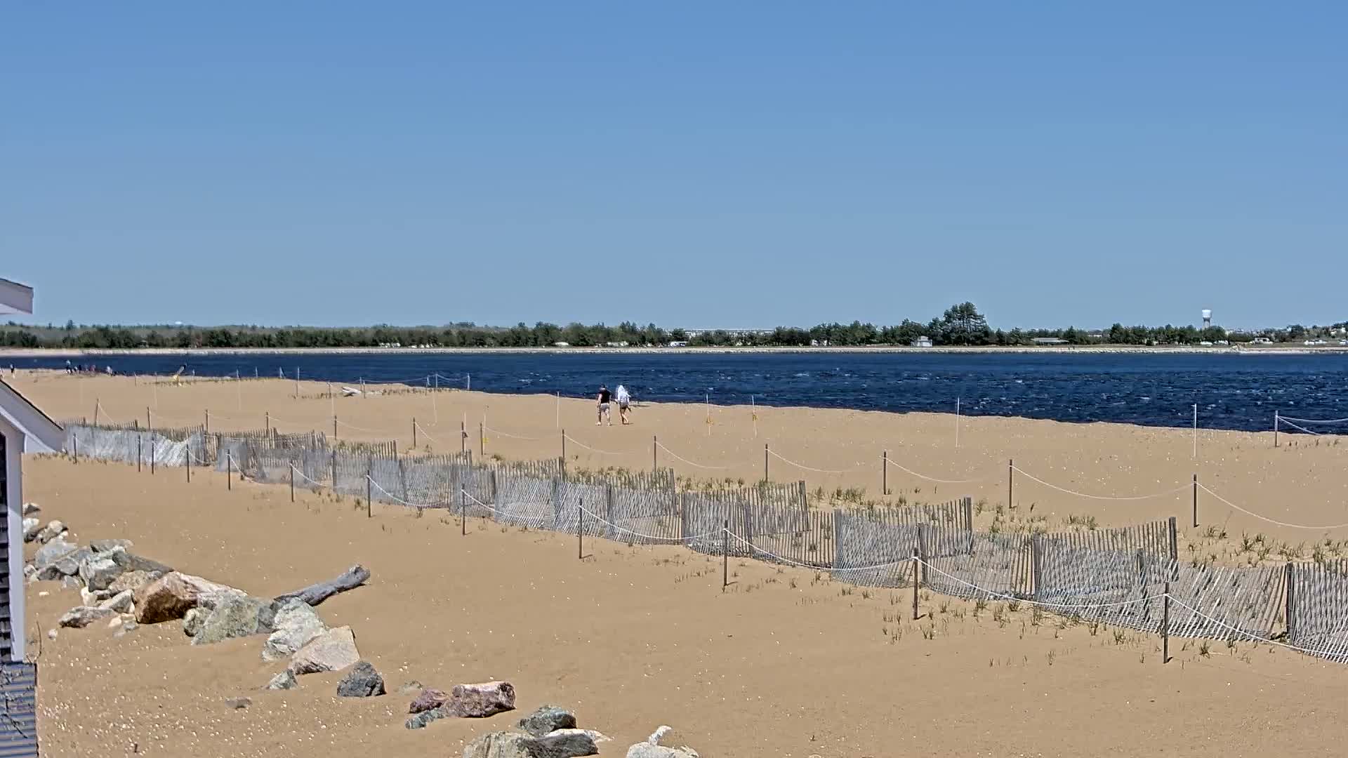 A sandy beach with a small fence protecting dune grasses, two people walking along the shoreline, and a choppy body of water under a clear blue sky.