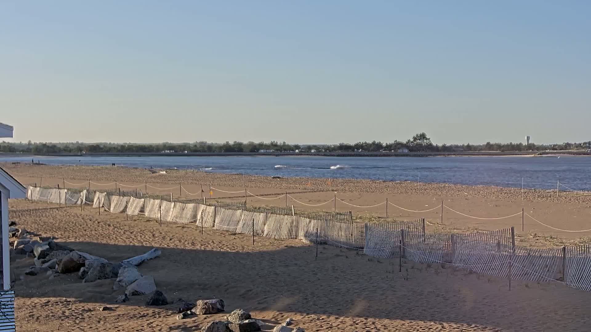 A sandy beach with a low wooden fence stretches along a calm body of water under a clear, sunny sky.