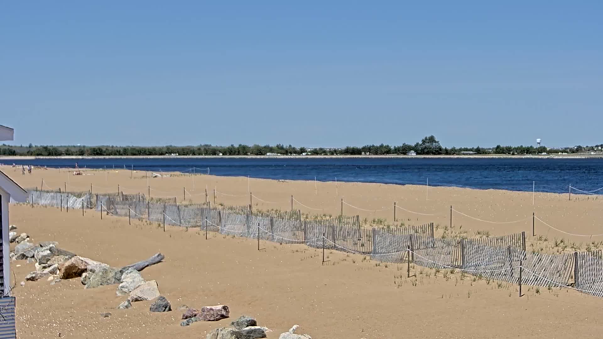 A sandy beach with a short wooden fence and a few people is visible under a clear blue sky, with calm water beyond.