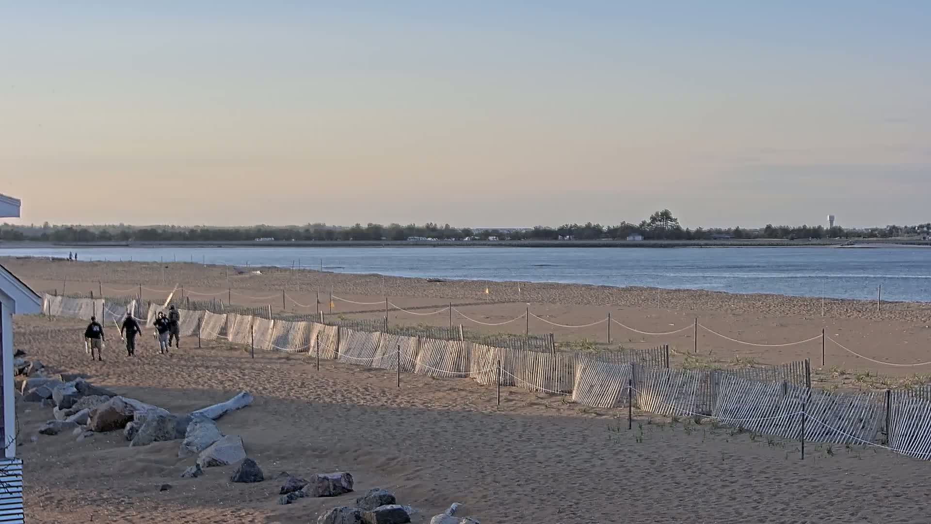 A sandy beach with a low wooden fence stretches towards a calm body of water under a clear, pale sky, and several people are walking along the beach.