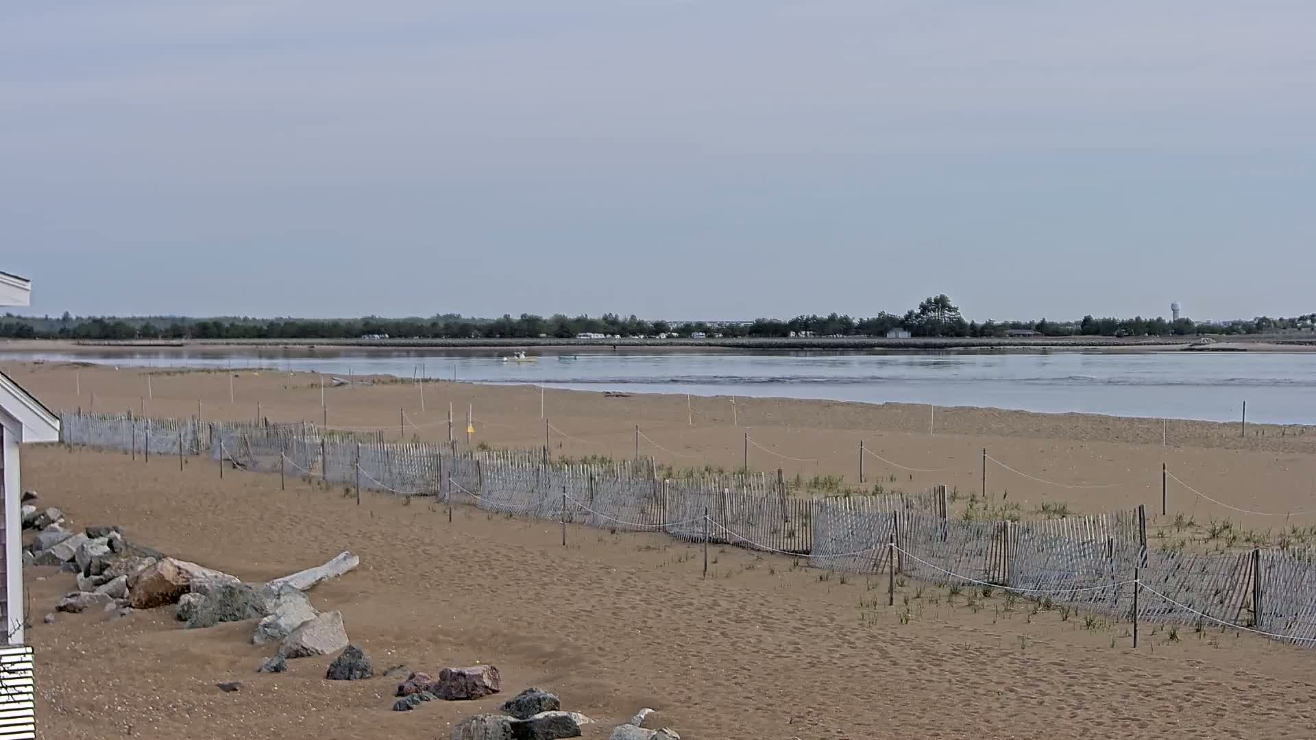 A sandy beach with a low wooden fence stretches towards a calm body of water under an overcast sky.