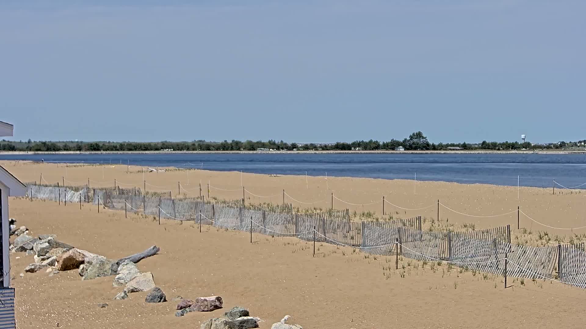 A sandy beach with a low wooden fence protecting young vegetation stretches toward a calm body of water under a clear blue sky.