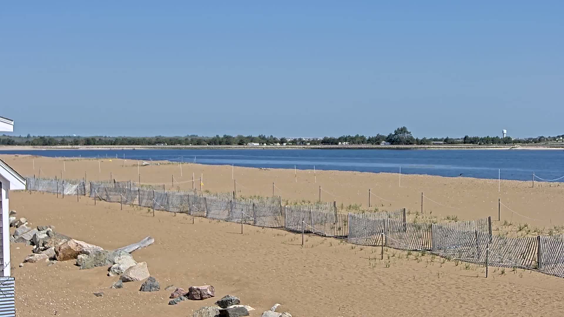 A sandy beach with a low wooden fence extends to a calm body of water under a clear blue sky.