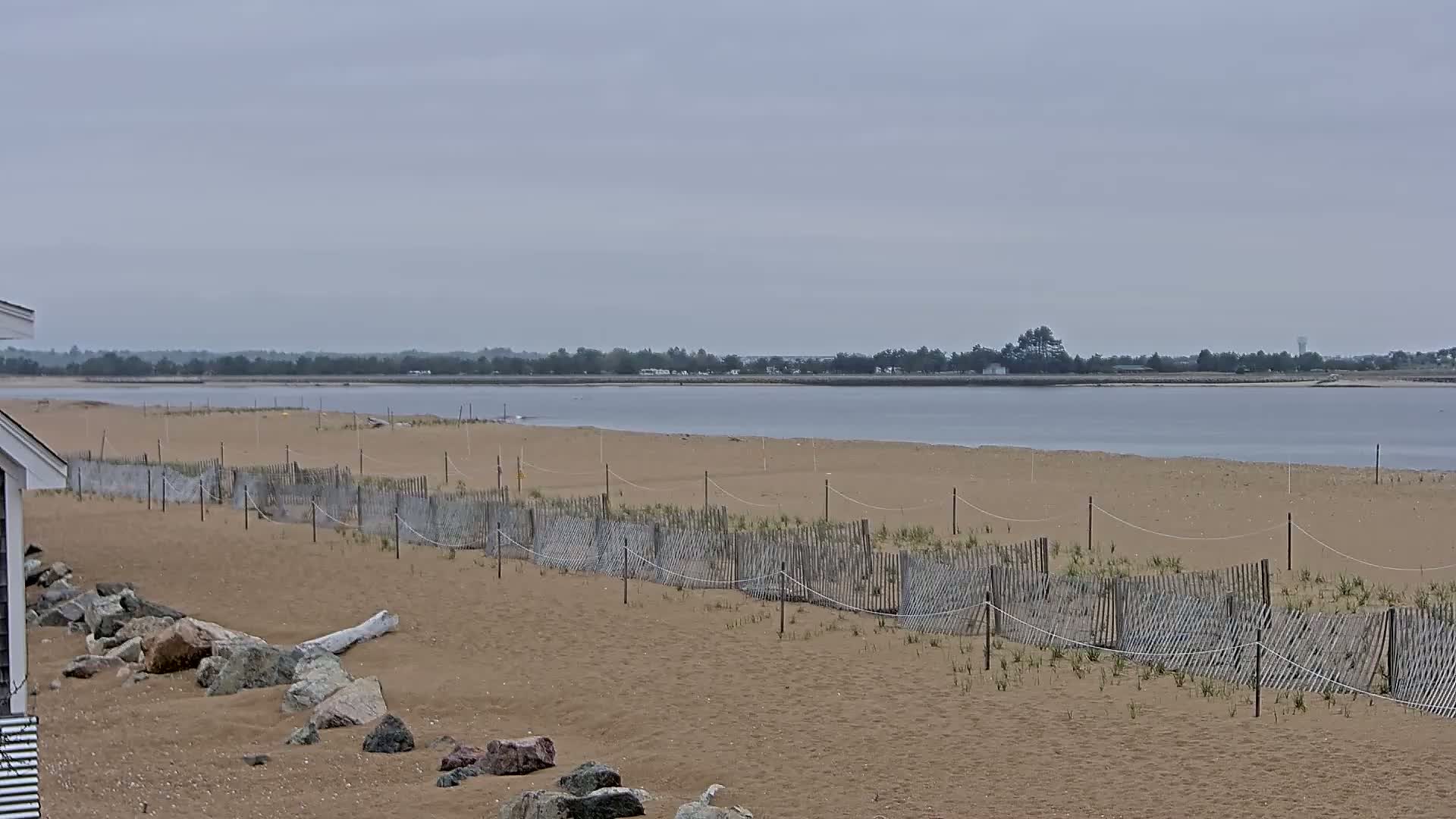 A sandy beach with a low wooden fence stretches toward a calm body of water under an overcast sky.