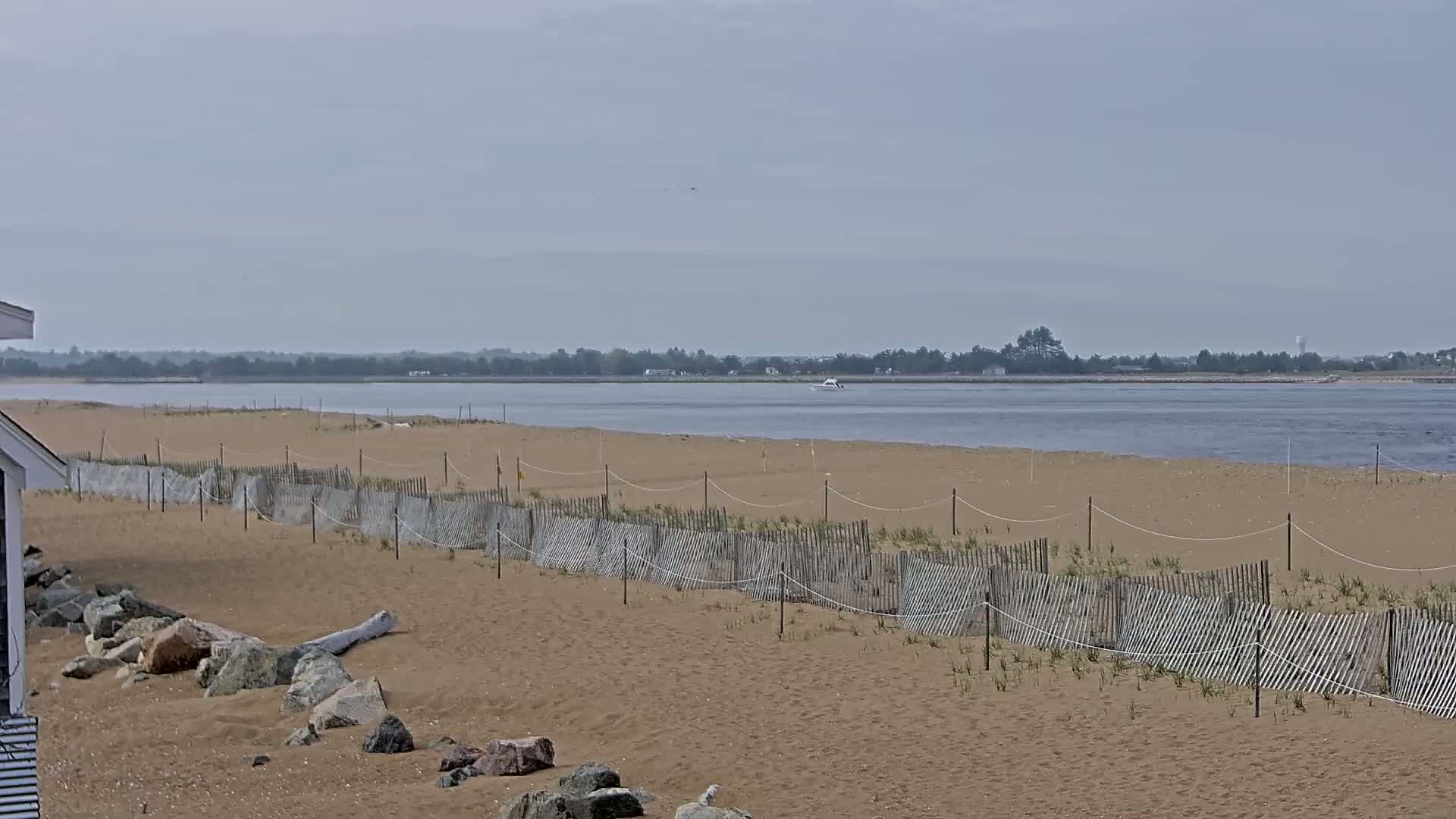 A sandy beach with a low wooden fence stretches to a calm body of water under an overcast sky.