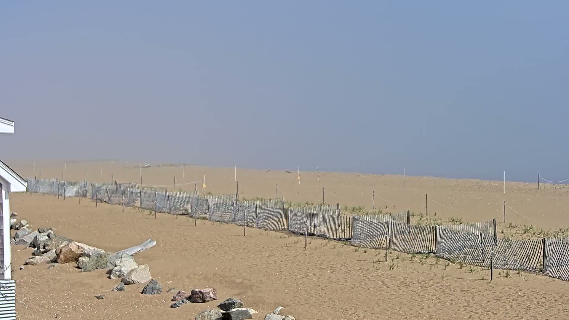 A hazy day reveals a sandy beach with a long, low fence running parallel to the water's edge, punctuated by rocks near a building's corner.