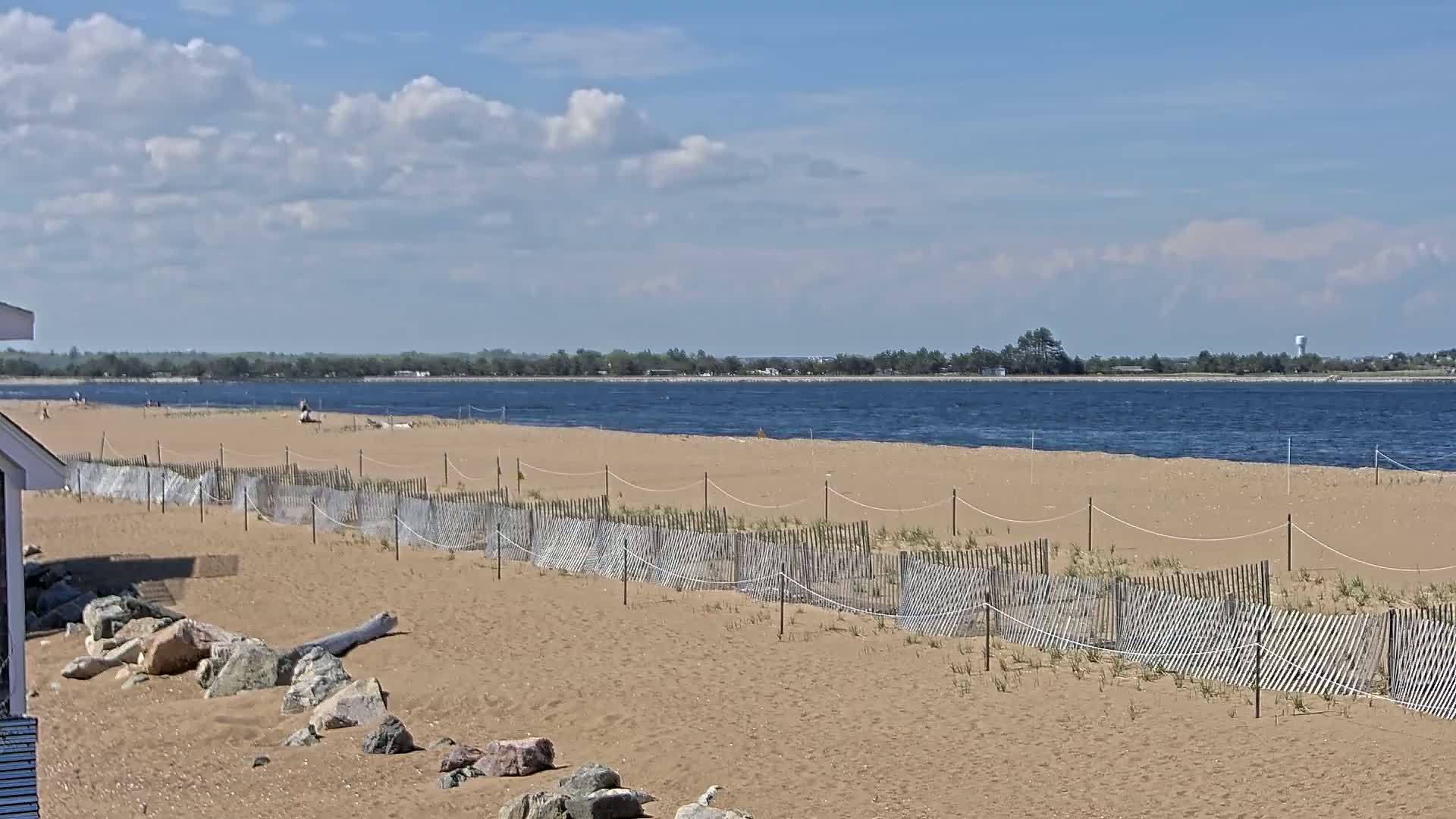 A sandy beach with a low wooden fence stretches along a calm, blue body of water under a partly cloudy sky.