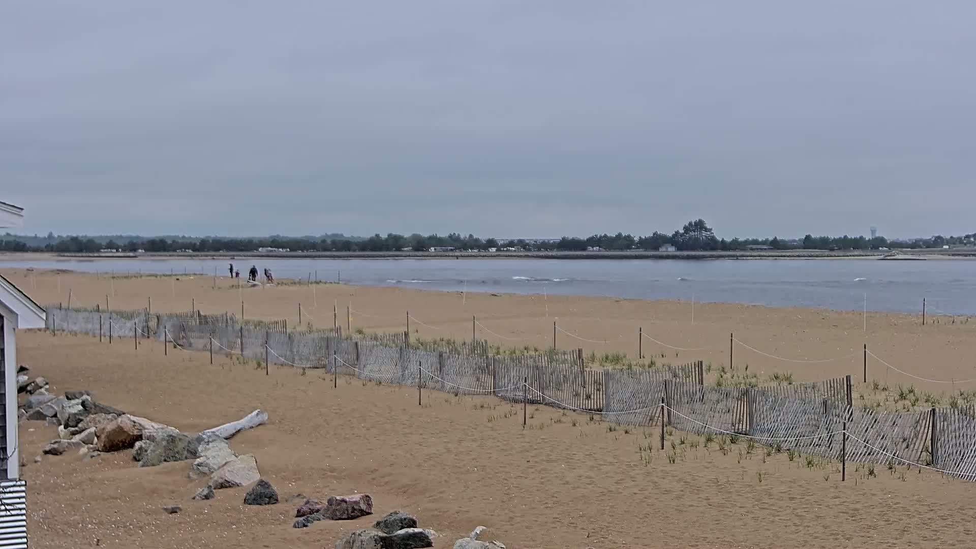 A sandy beach with a short wooden fence, a few people in the distance, and calm water under an overcast sky.