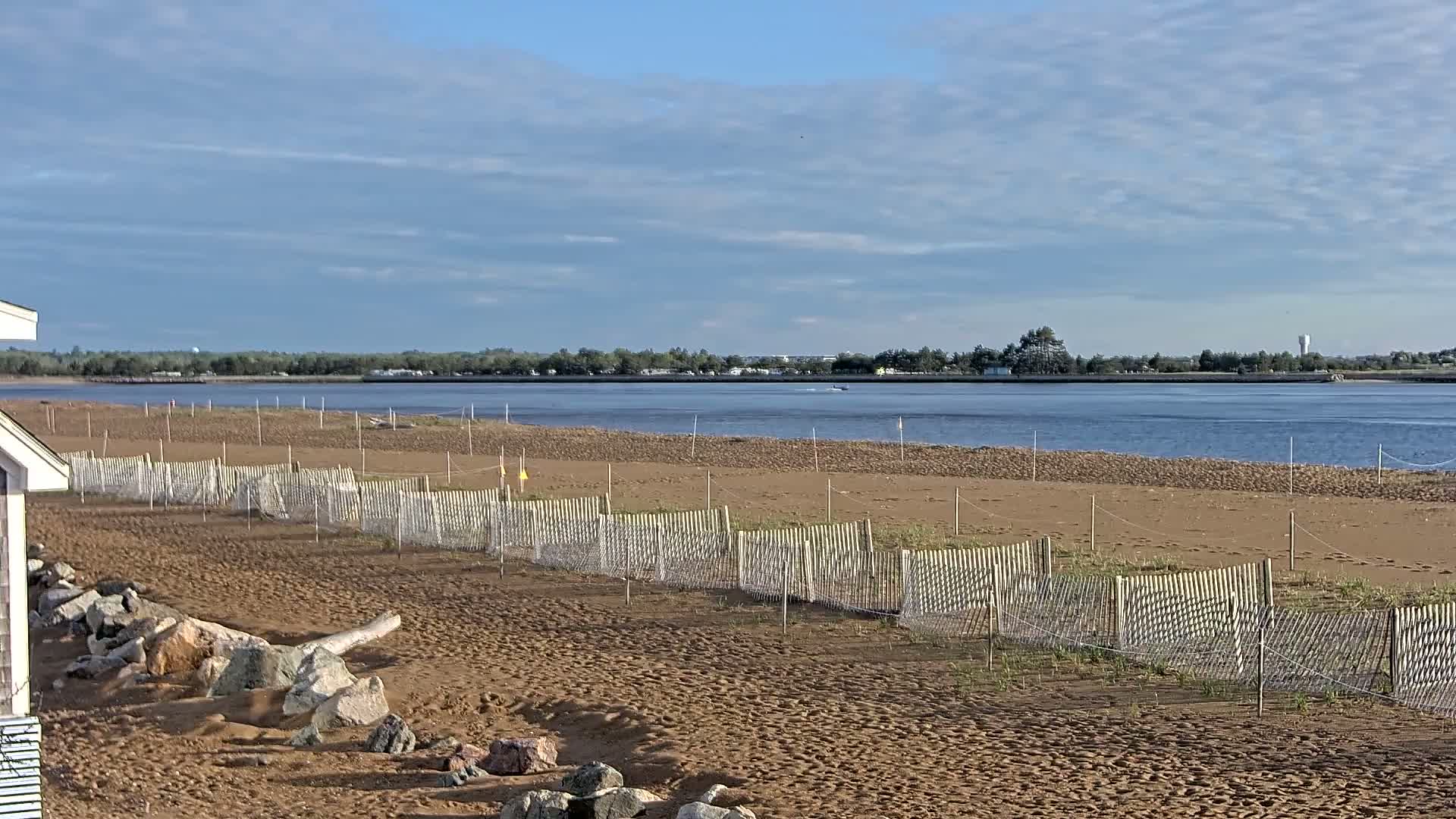 A sandy beach with a low wooden fence stretches towards a calm body of water under a partly cloudy sky.