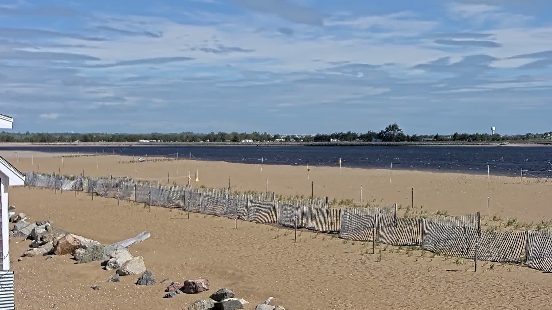 A sandy beach with a wooden fence stretches toward a calm body of water under a partly cloudy sky.