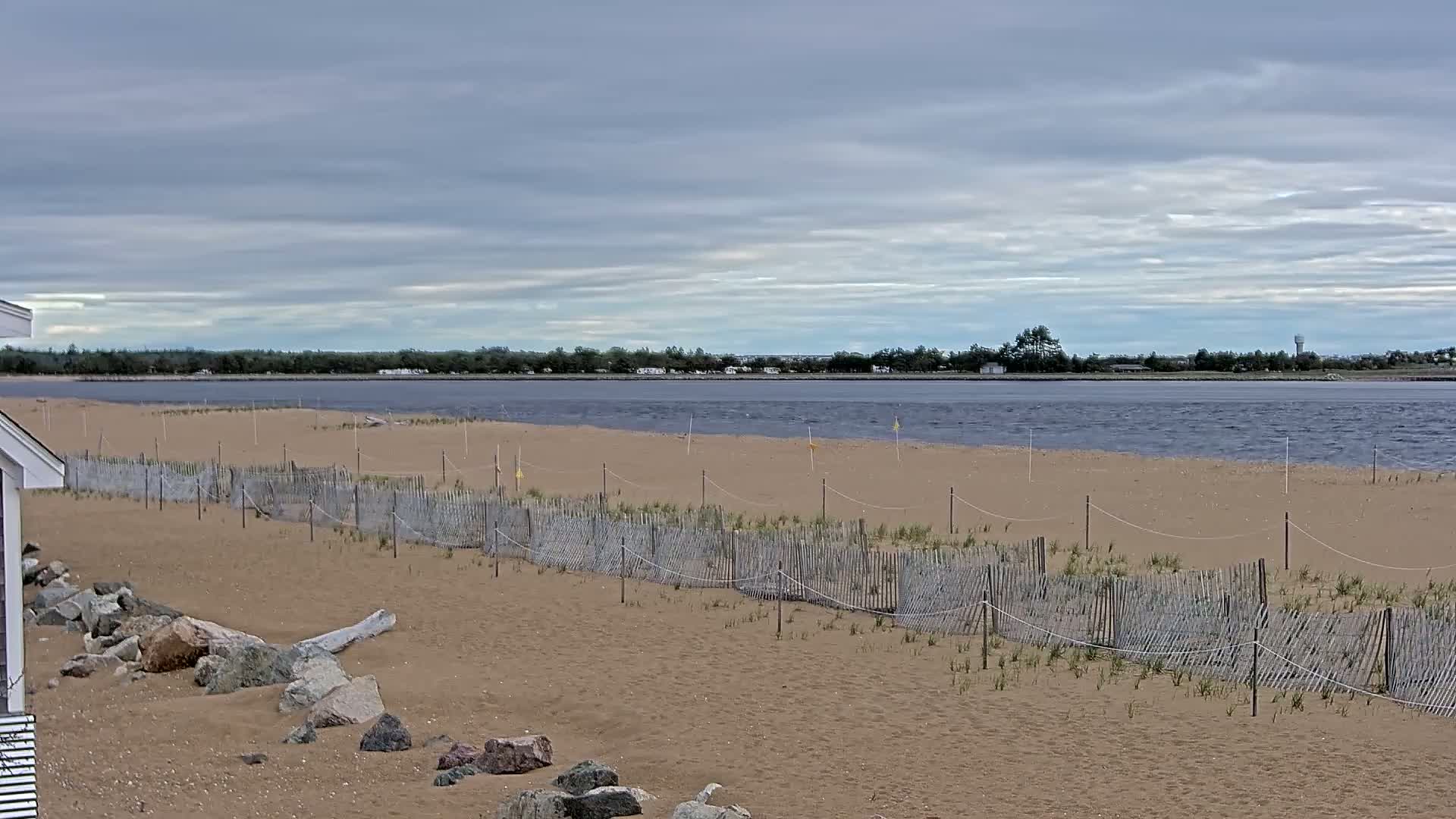 A sandy beach with a low wooden fence stretches along a calm, gray-water bay under a cloudy sky.