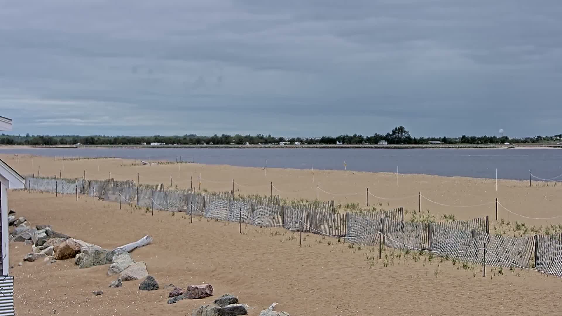 A sandy beach with a low wooden fence stretches towards a calm, purplish-gray body of water under an overcast sky.