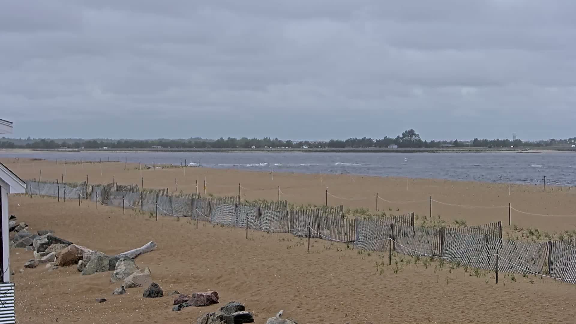A sandy beach with a low wooden fence stretches towards a calm, gray body of water under an overcast sky.