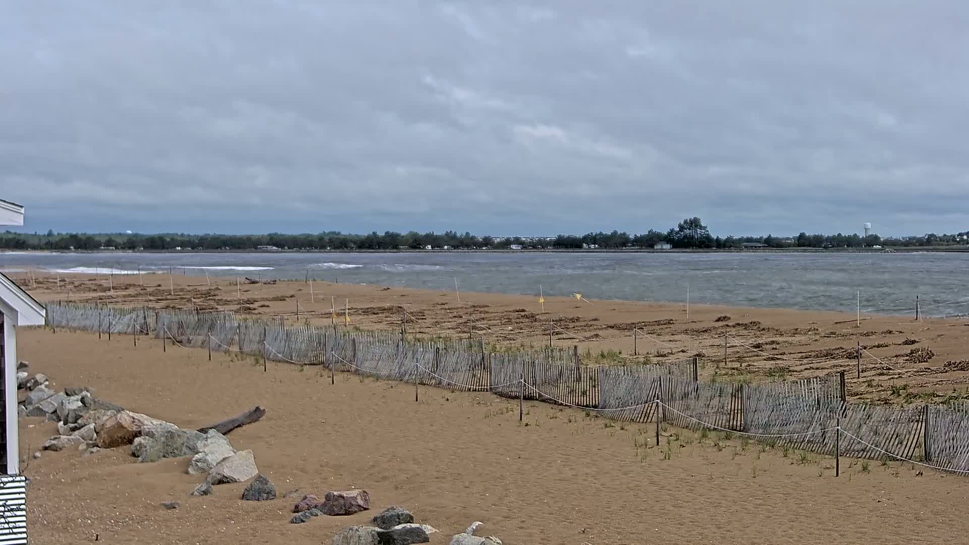 A sandy beach with a wooden fence, partially obscured by windblown debris, stretches toward a choppy body of water under a cloudy sky.