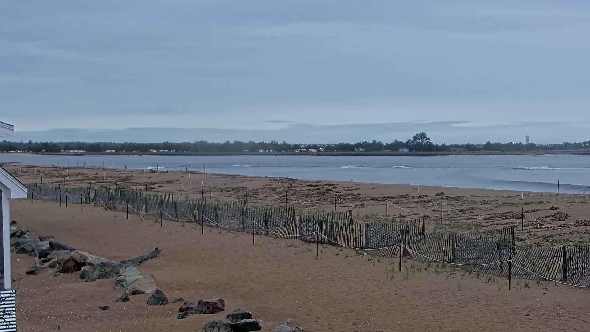 A sandy beach with a short wooden fence stretches towards a calm, grey-sky ocean under an overcast sky.