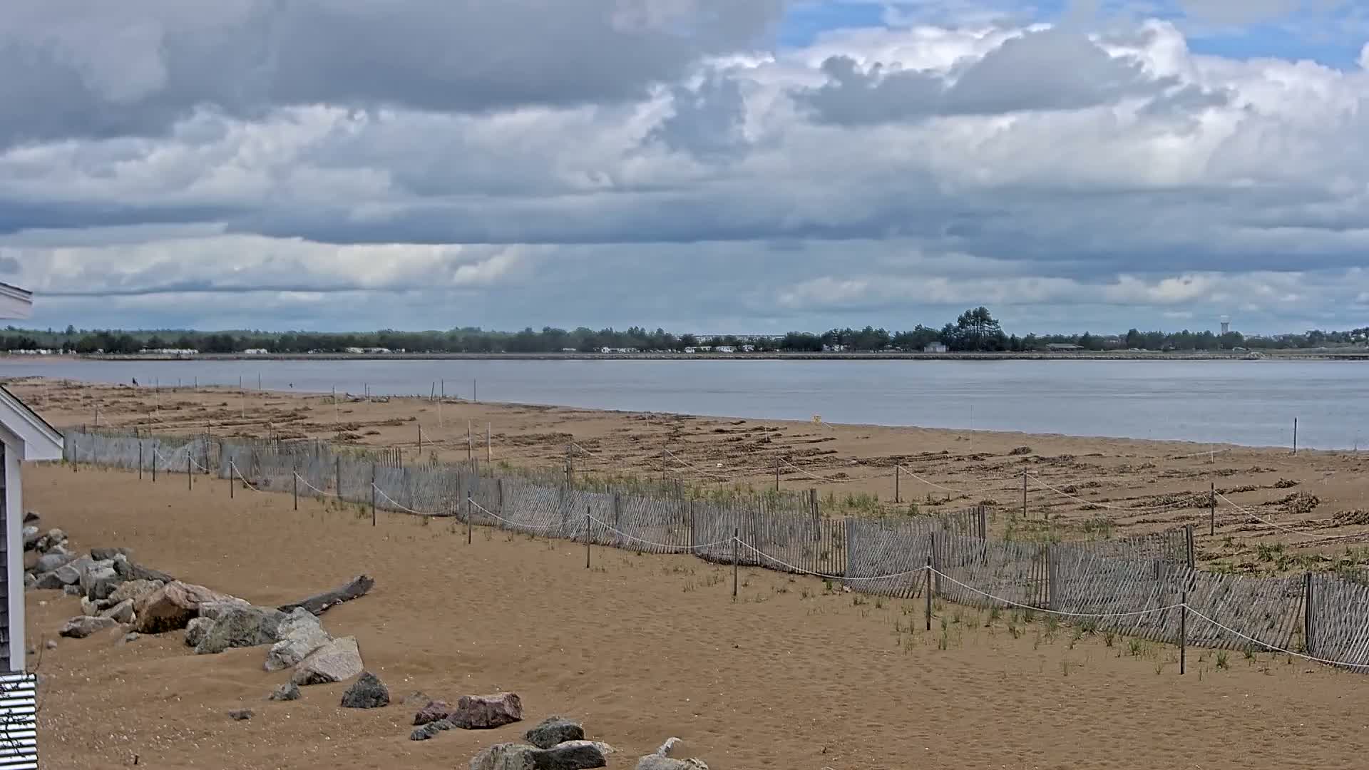 A sandy beach with a wooden fence stretches towards a calm body of water under a cloudy sky.