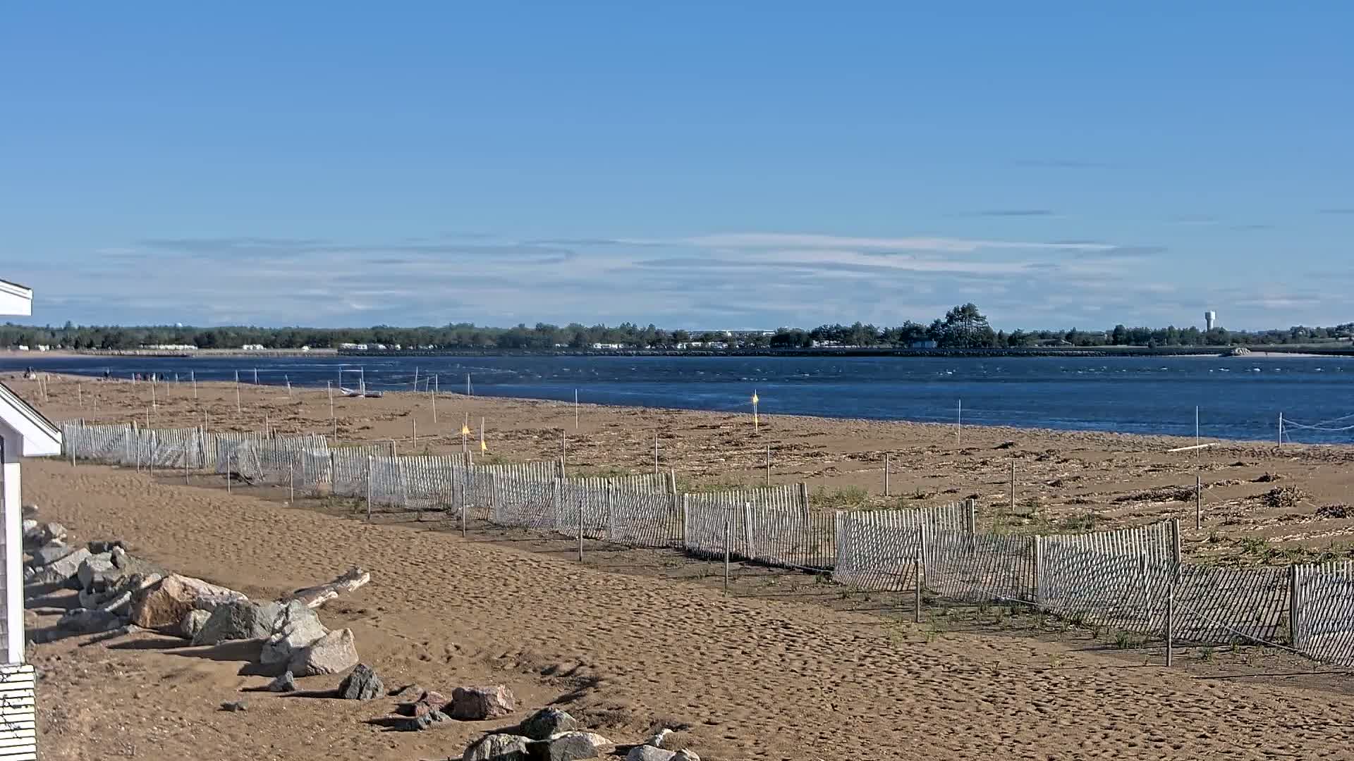 A sandy beach with a wooden fence stretches along a calm body of water under a mostly clear blue sky.