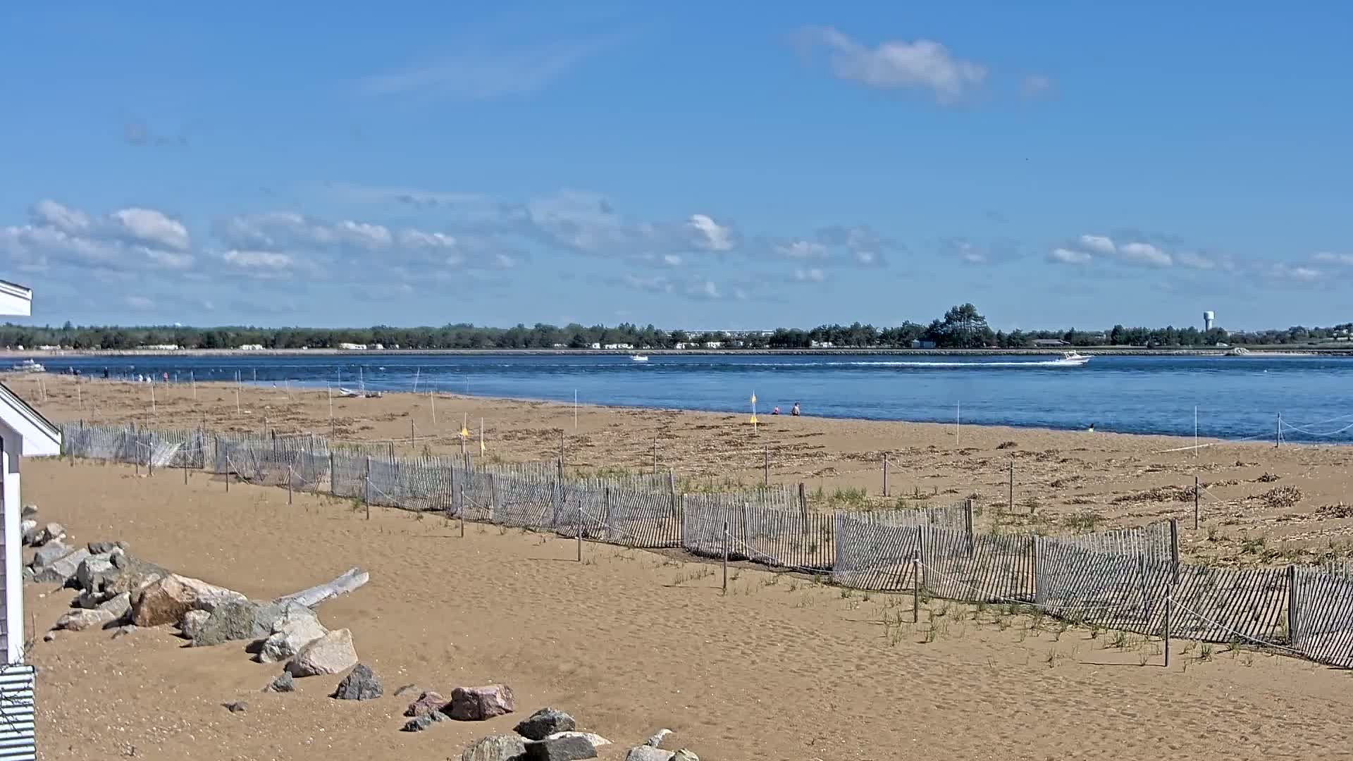 A sandy beach with a wooden fence and a few people is visible, overlooking a calm body of water under a mostly sunny sky with scattered clouds.