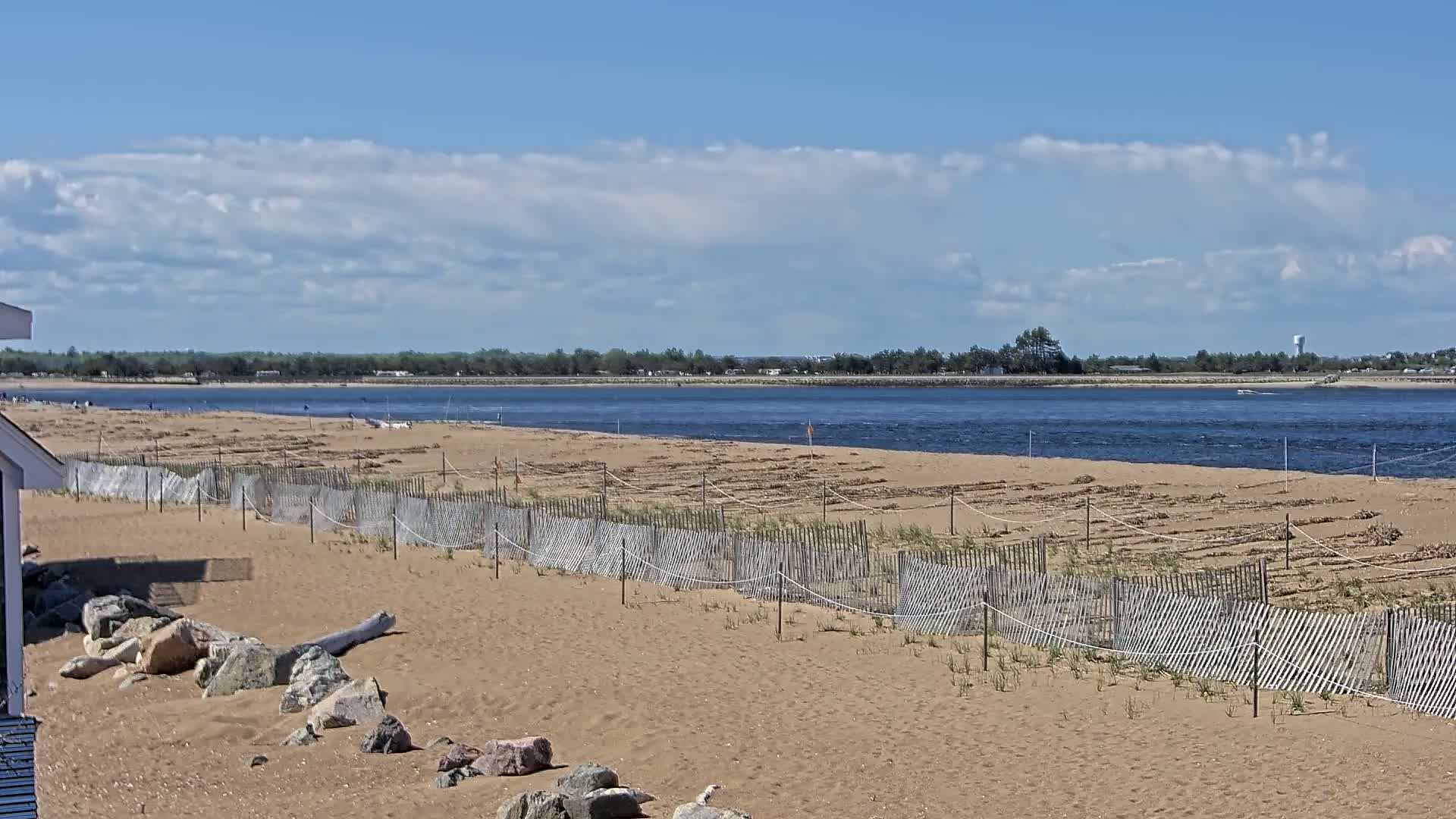 A sandy beach with a low wooden fence stretches along a calm body of water under a partly cloudy sky.