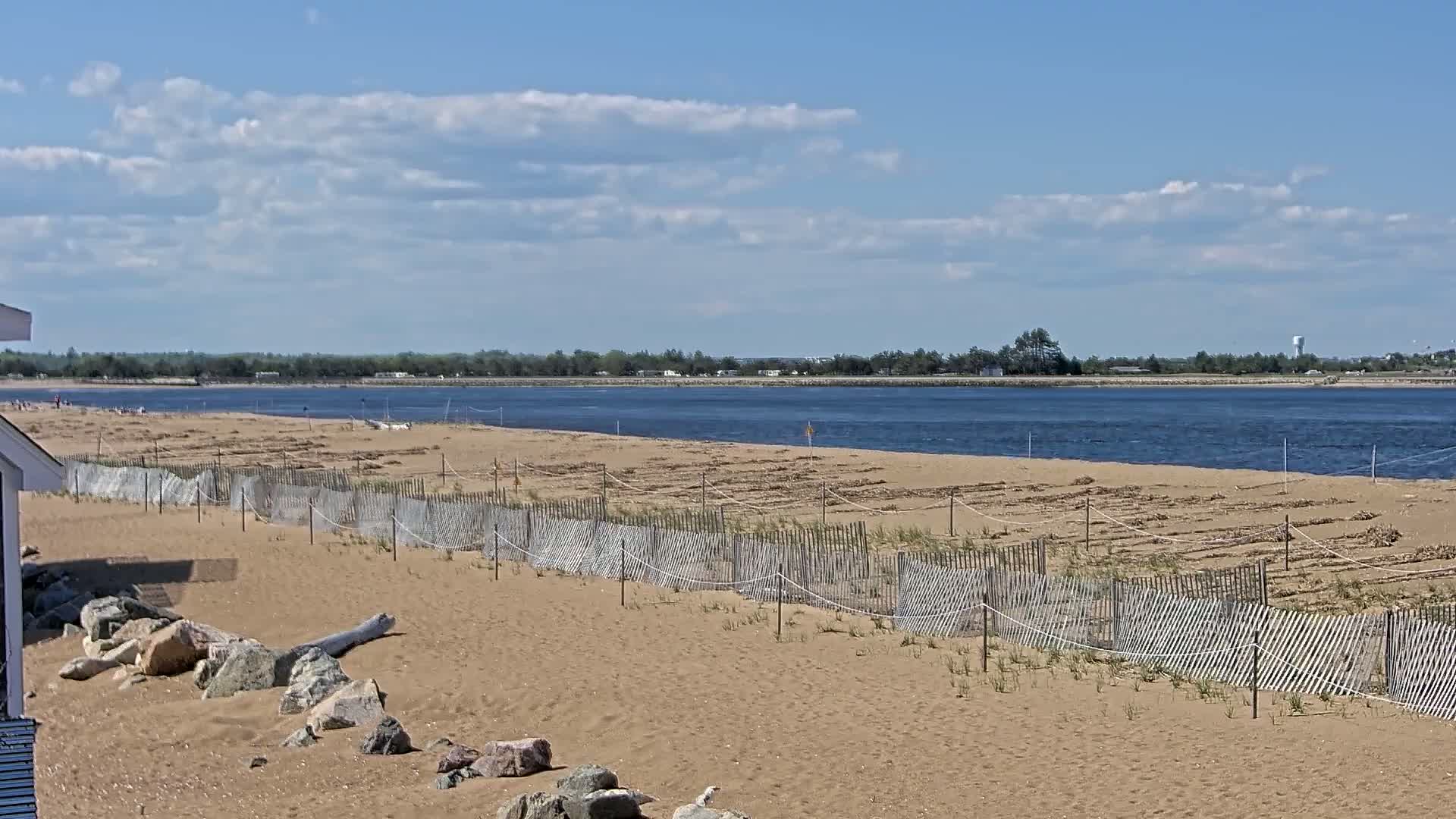 A sandy beach with a short wooden fence protecting rows of vegetation, stretches along a calm body of water under a partly cloudy sky.