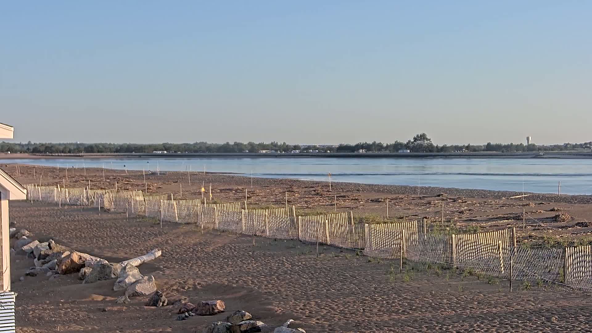 A sandy beach with a low wooden fence stretches along a calm river under a clear, bright sky.