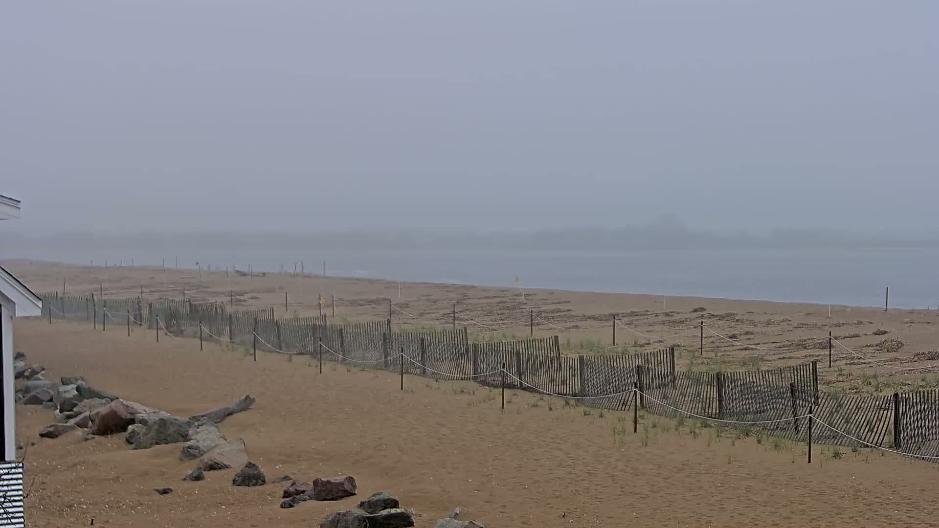 A foggy beach scene shows a sandy shore with a low wooden fence running parallel to the water's edge.