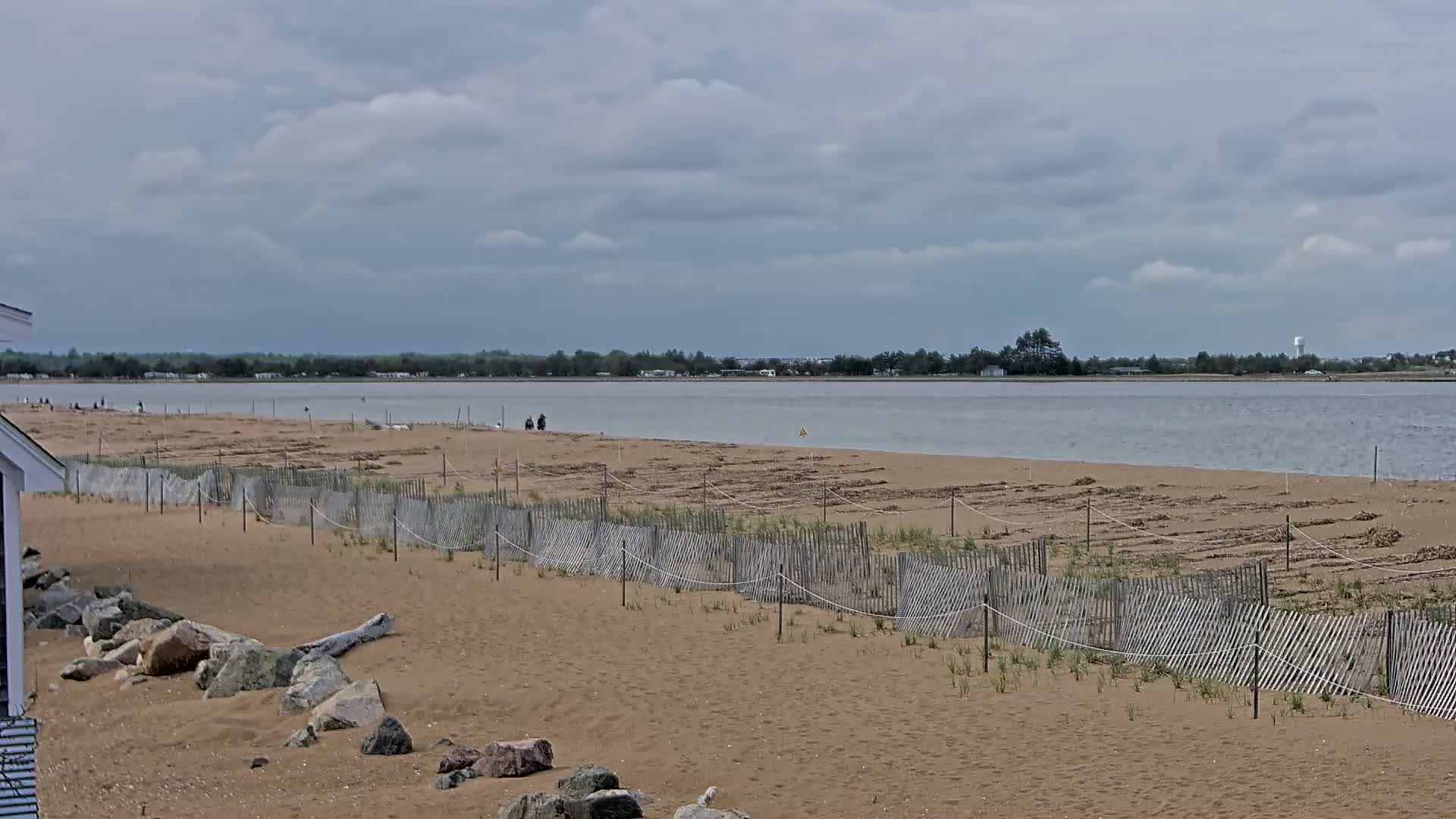 A sandy beach with a wooden fence and a calm body of water under a cloudy sky is visible.